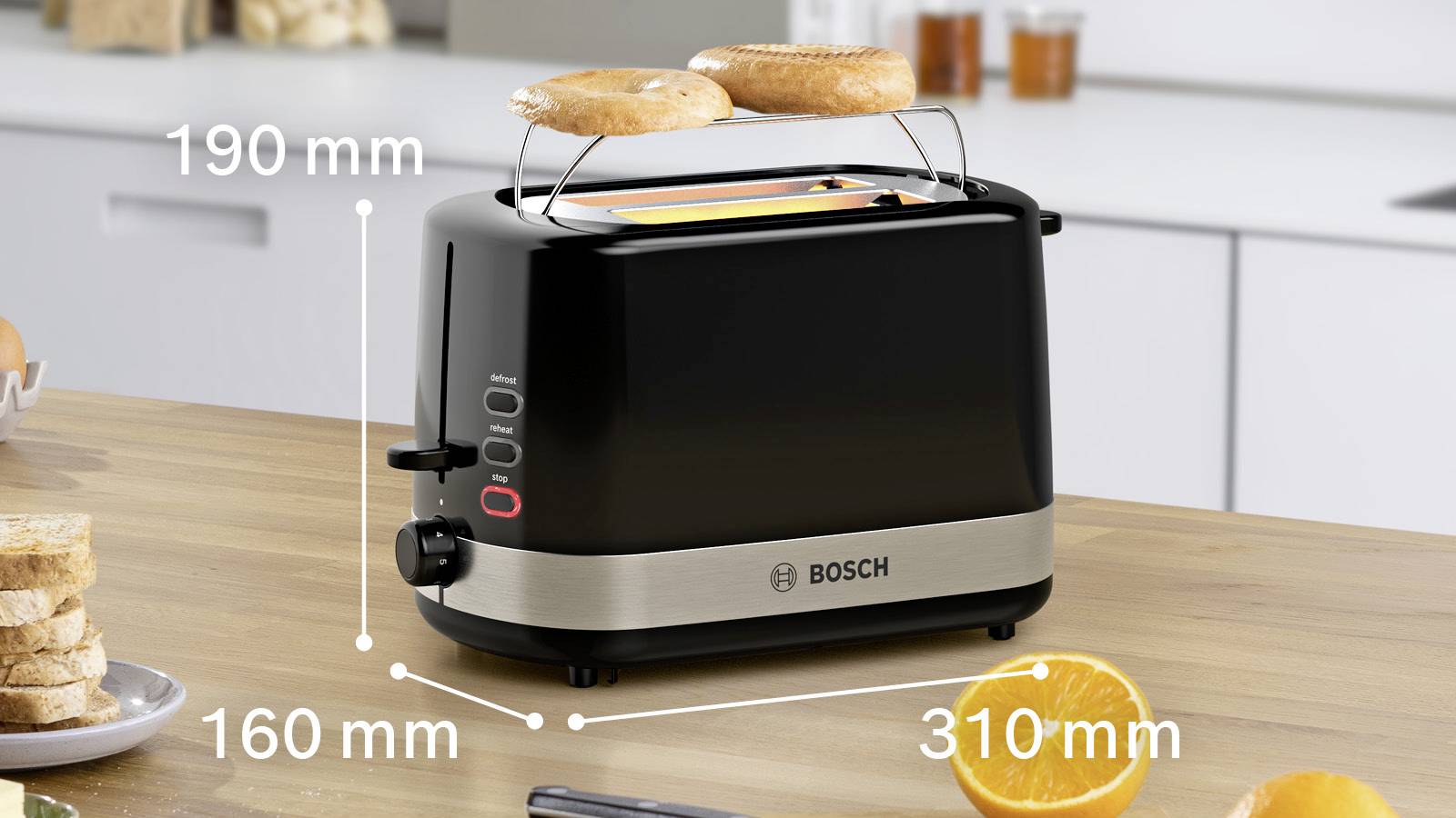 Ein schwarzer Toaster auf einer Küchenarbeitsfläche röstet zwei Bagel. Maße: Höhe 190 mm, Tiefe 160 mm, Breite 310 mm. Kleines Messer und Orangen im Vordergrund.