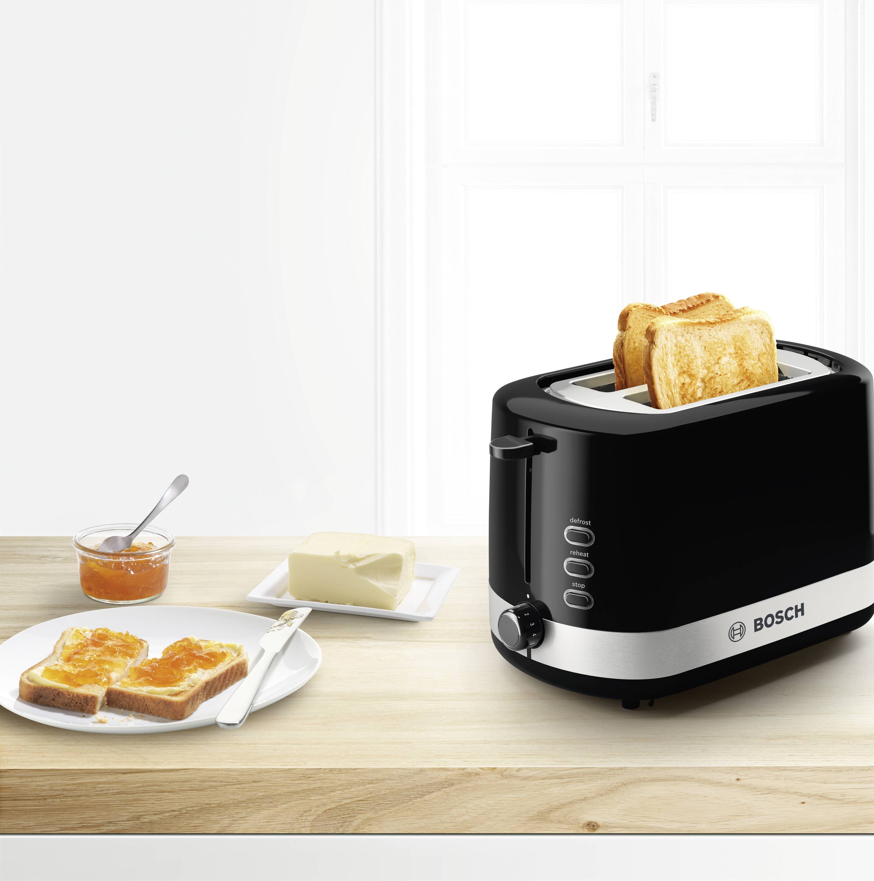 Ein schwarzer Toaster röstet zwei Scheiben Toast. Daneben ein Teller mit Marmeladentoast und ein Stück Butter auf einem Holzbrett. Im Hintergrund ein Fenster.
