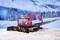 Eine Pistenraupe räumt Schnee in einer verschneiten Berglandschaft mit bewaldeten Hügeln und Bergen im Hintergrund.