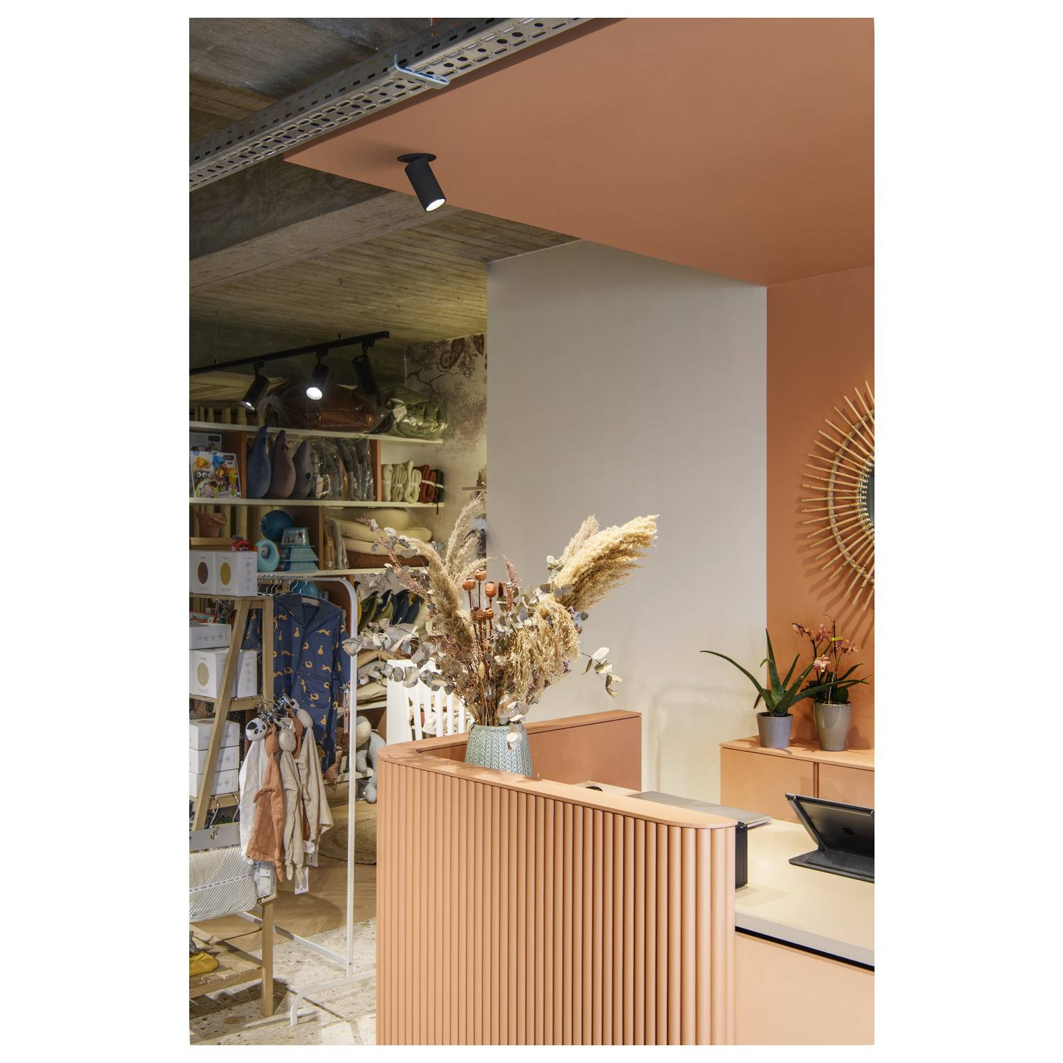 Ein stilvoller Ladenbereich mit orangener Theke. Trockenblumen in einer Vase als Dekoration. Regale mit Kleidung und Accessoires im Hintergrund.