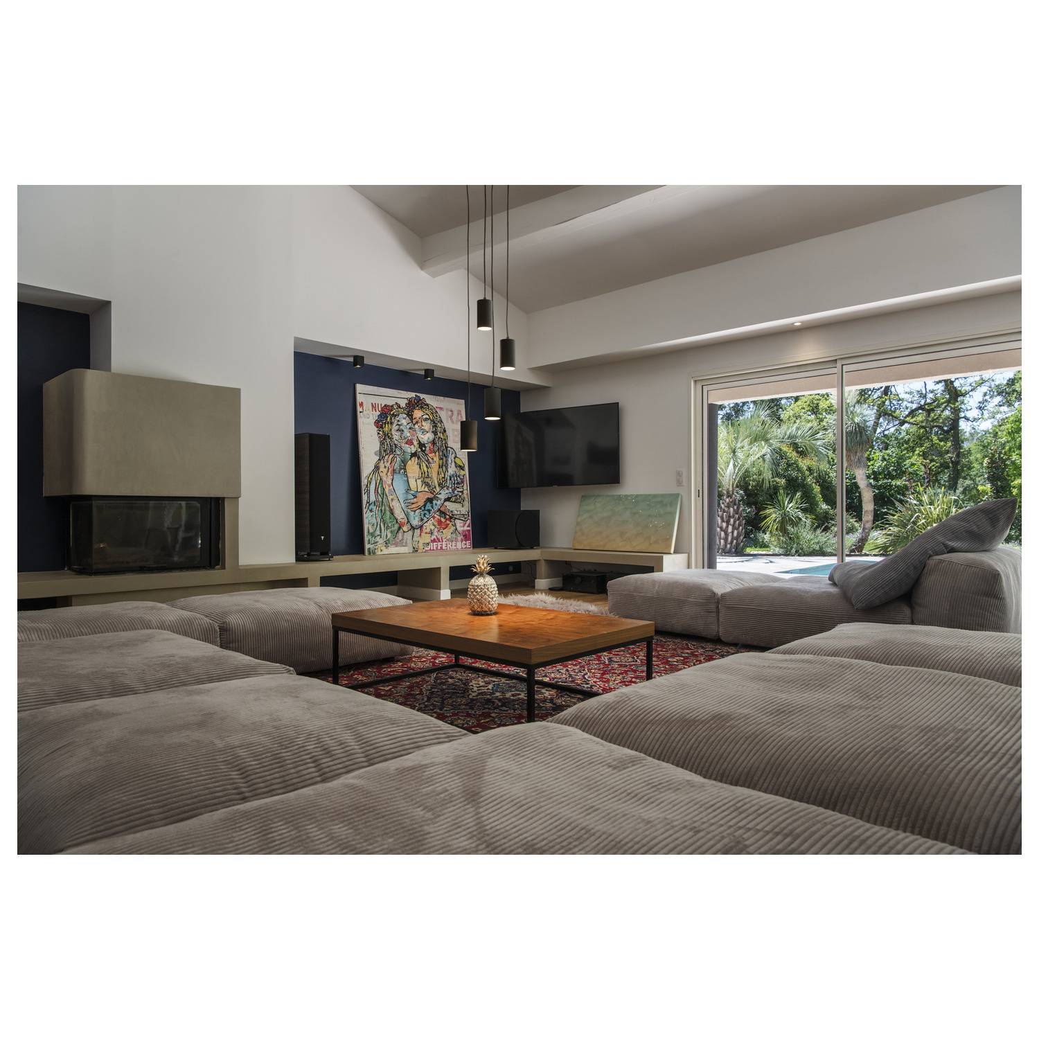 Gemütliches Wohnzimmer mit grauem Sofa, Holztisch und großem Fenster. An der Wand hängt ein buntes Gemälde. Draußen sind Bäume sichtbar.