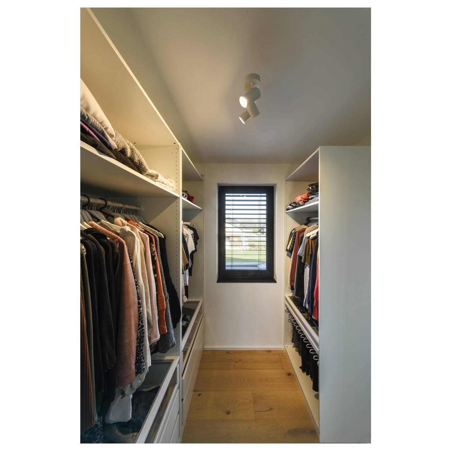 Ein begehbarer Kleiderschrank mit hölzernem Boden, Regalen voller Kleidung und einem Fenster mit Jalousien am Ende des Raums.
