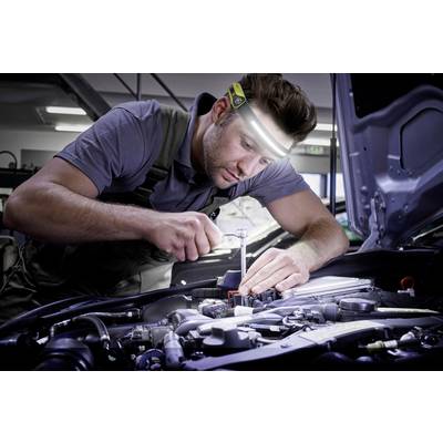 Ein Mechaniker, der konzentriert an einem Auto-Motor arbeitet, trägt eine Stirnlampe. Im Hintergrund sind Werkzeuge und Werkstattbeleuchtung sichtbar.