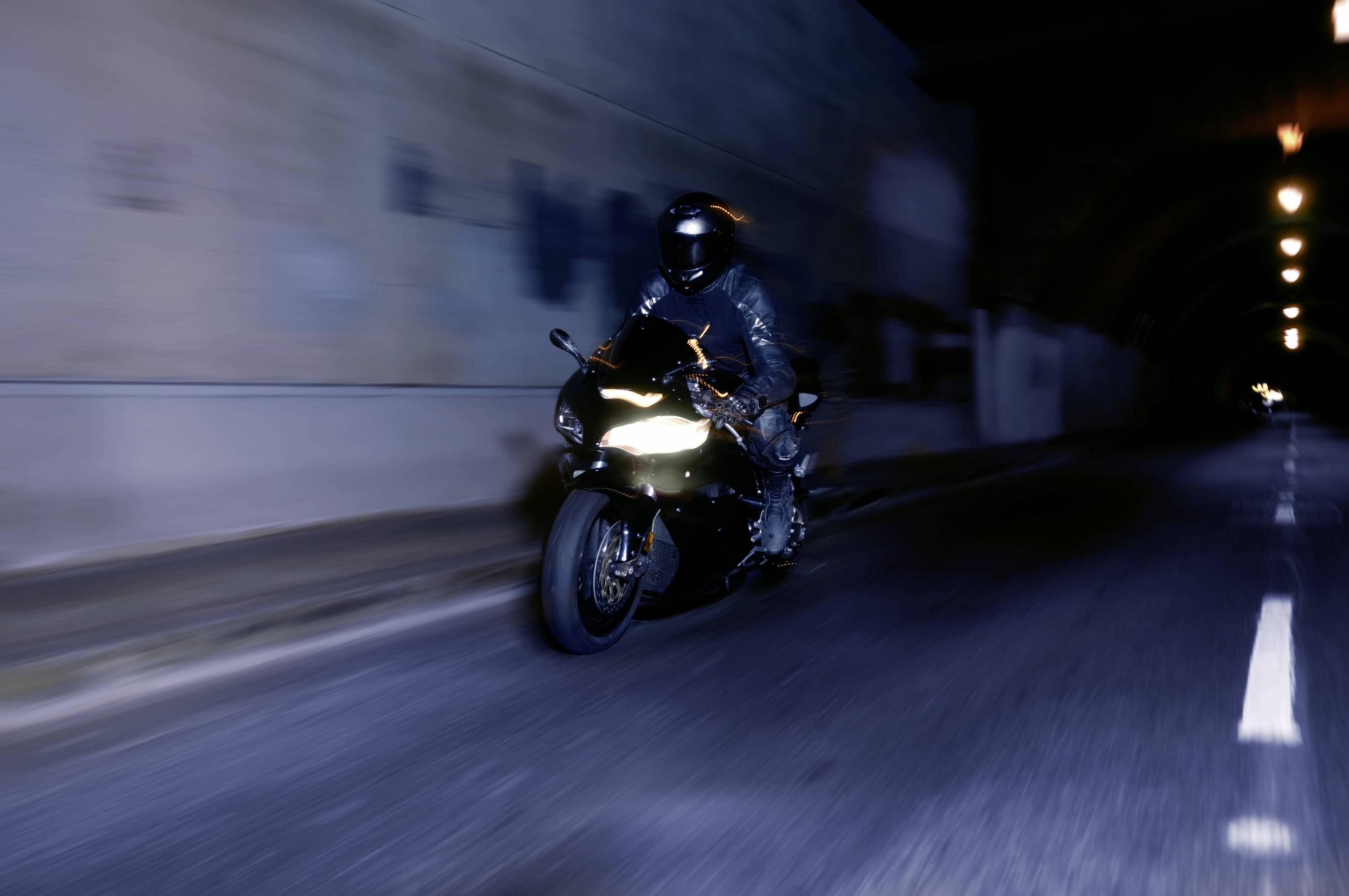 Motorradfahrer in schwarzer Schutzkleidung fährt bei Nacht durch einen beleuchteten Tunnel. Geschwindigkeit und Bewegung sind deutlich sichtbar.