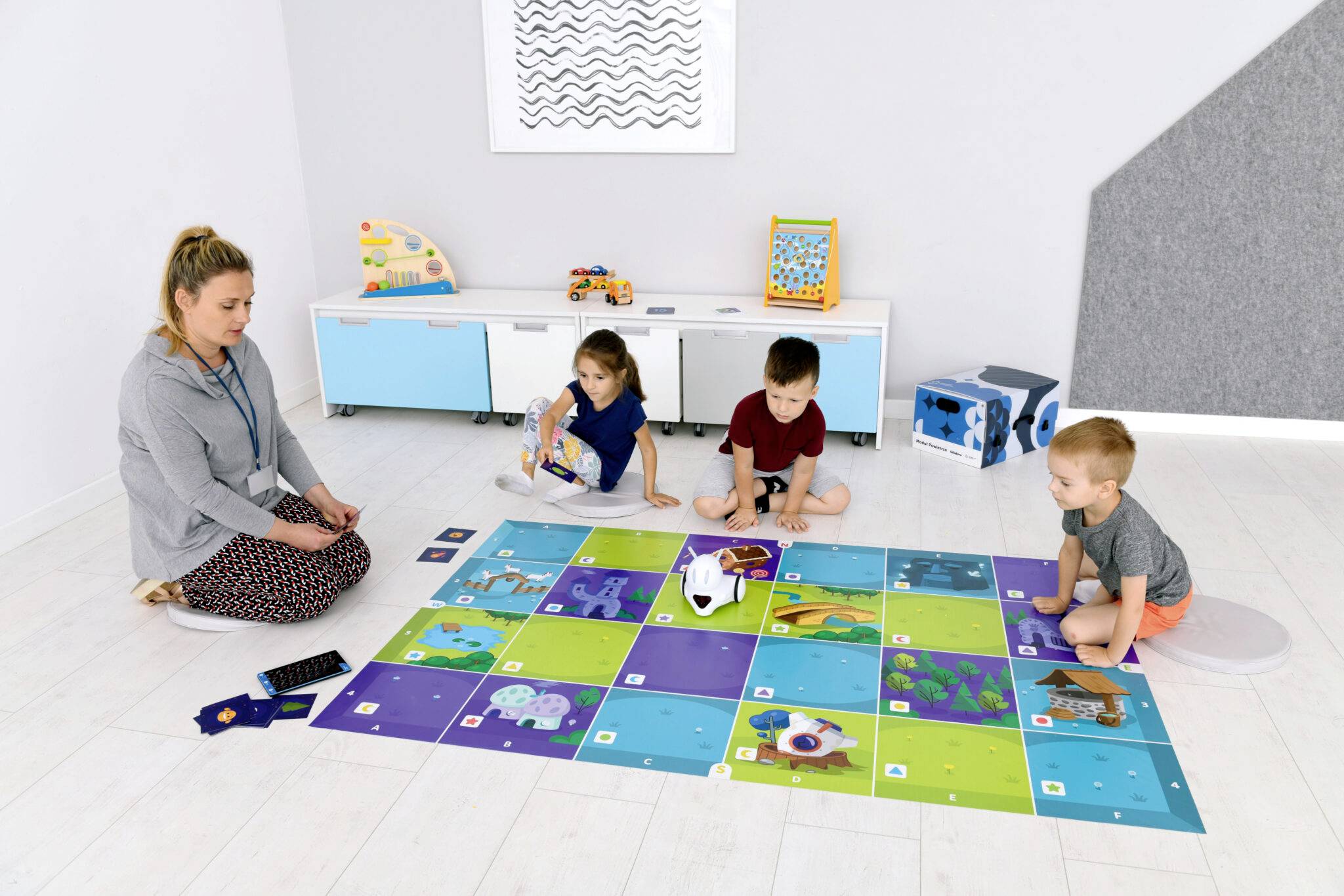 Eine Erzieherin zeigt drei Kindern, wie man mit einem Roboter auf einer bunten Lernmatte aus Kartons Teile des Raums erkundet.