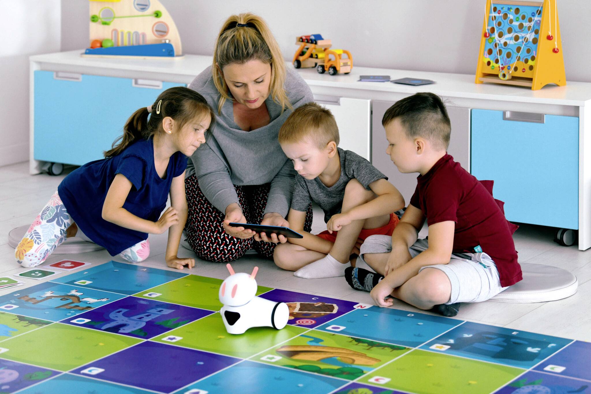 Eine Erzieherin und drei Kinder sitzen auf dem Boden und schauen auf ein Tablet. Vor ihnen bewegt sich ein kleiner Roboter auf bunten Quadraten.