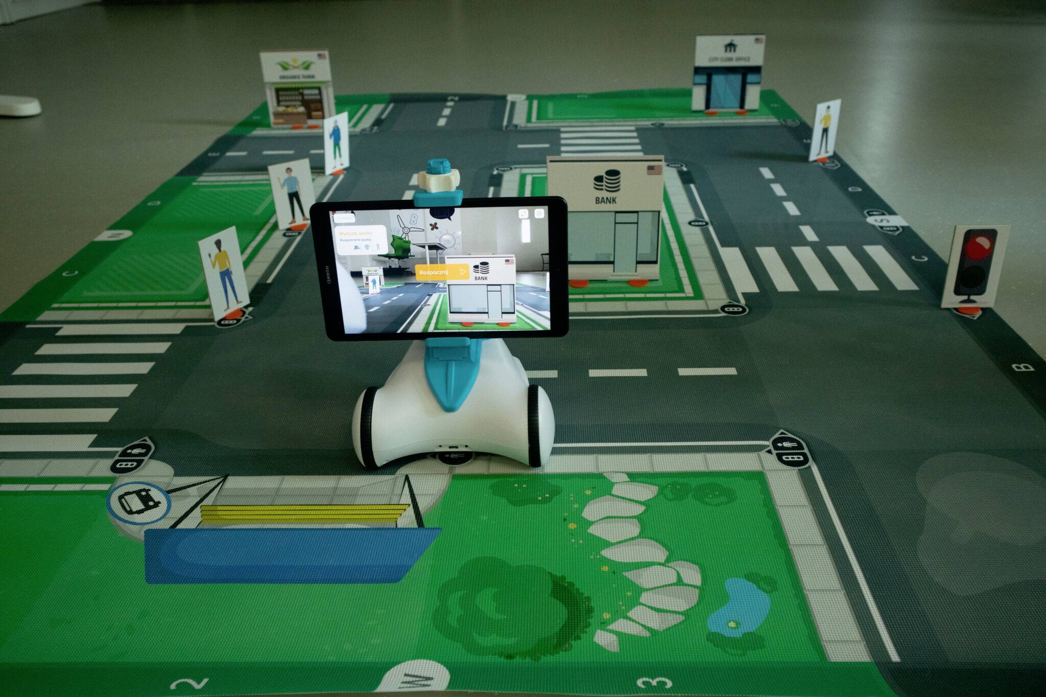 Ein Modell einer Stadt mit Straßen, Zebrastreifen und Gebäuden. Ein Roboter mit Tablet zeigt eine App an, um das Modell zu navigieren.