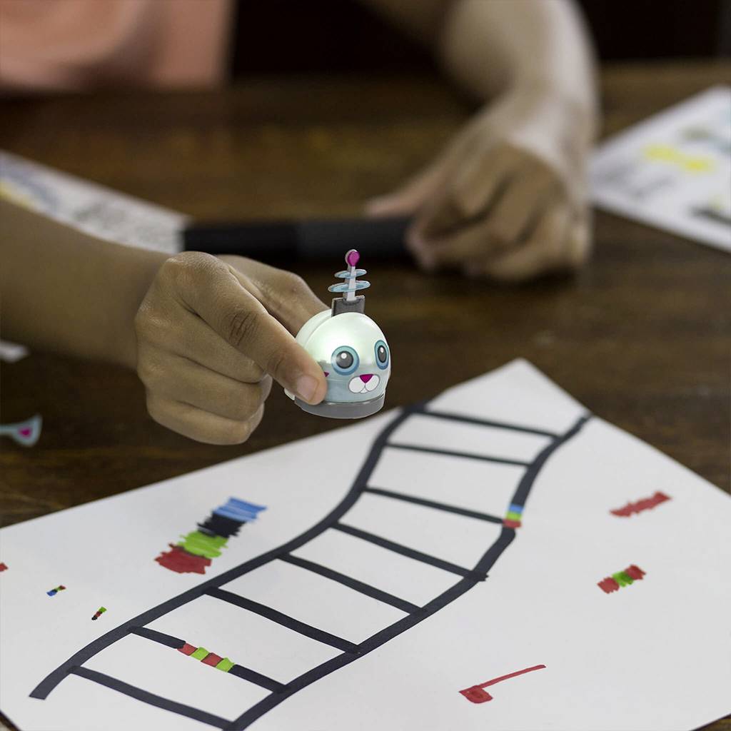 Ein Kind steuert einen kleinen Roboter über eine weiße Papierstraße mit Linien und bunten Markierungen. Der Roboter ist rund und lächelt.