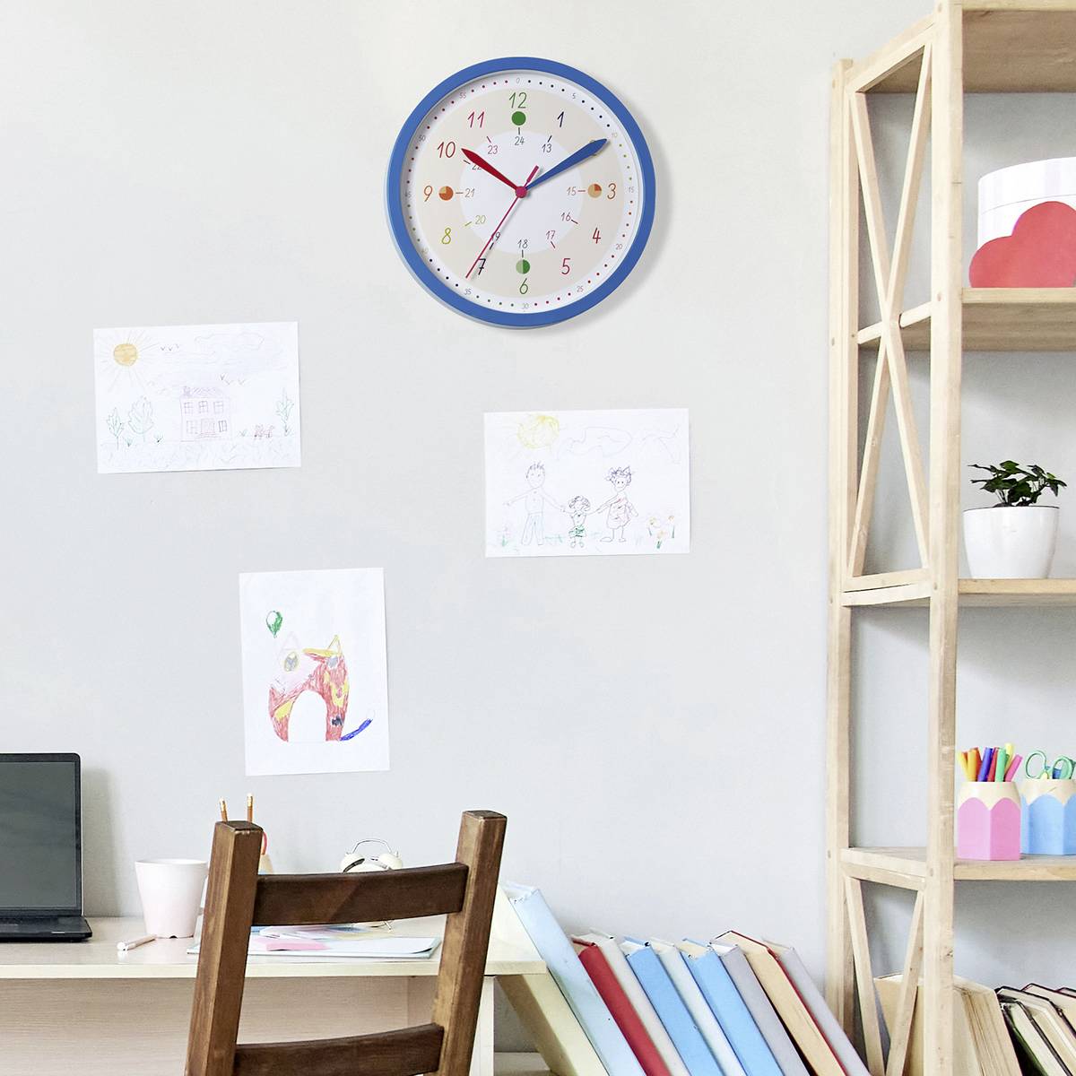 Ein Büro mit einem Schreibtisch, auf dem ein Laptop steht. An der Wand hängt eine Uhr. Bücher und Zeichnungen sind sichtbar.