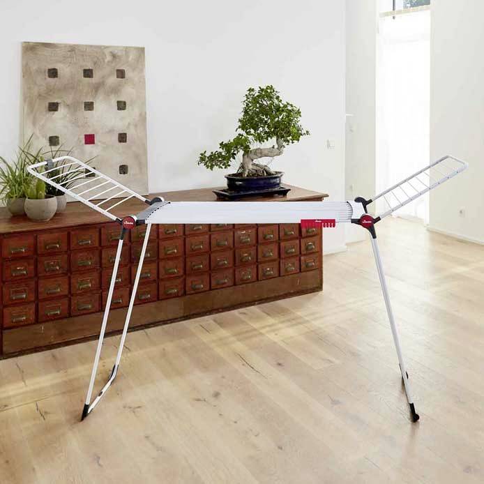 Ein zusammenklappbarer Wäscheständer steht in einem Raum mit Holzboden. Im Hintergrund befindet sich ein Sideboard mit mehreren Schubladen und einer Pflanze darauf.