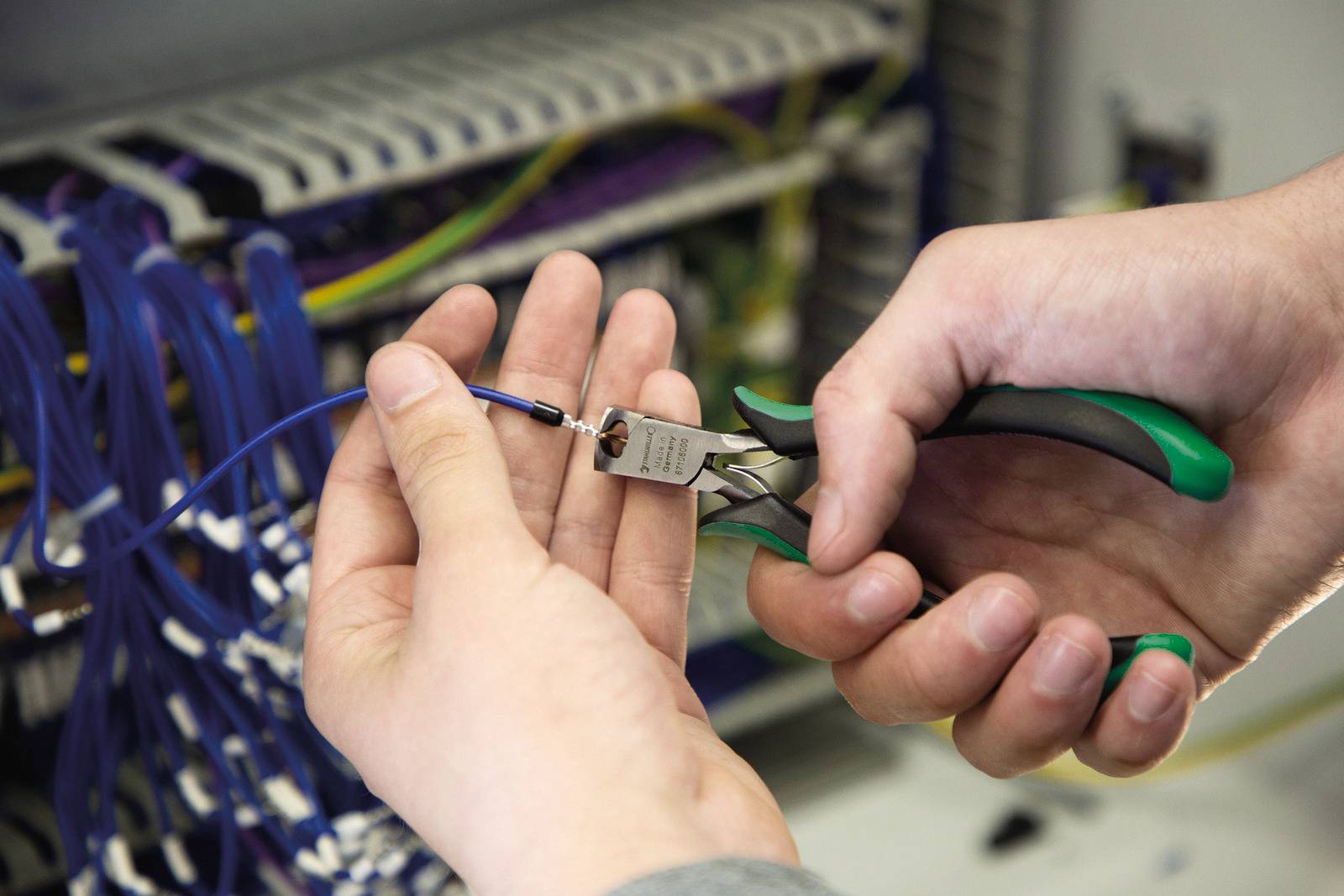 Hände halten eine Zange und ein Kabel vor einem geöffneten Schaltschrank. Die Szene zeigt elektrische Arbeiten oder Reparaturen.