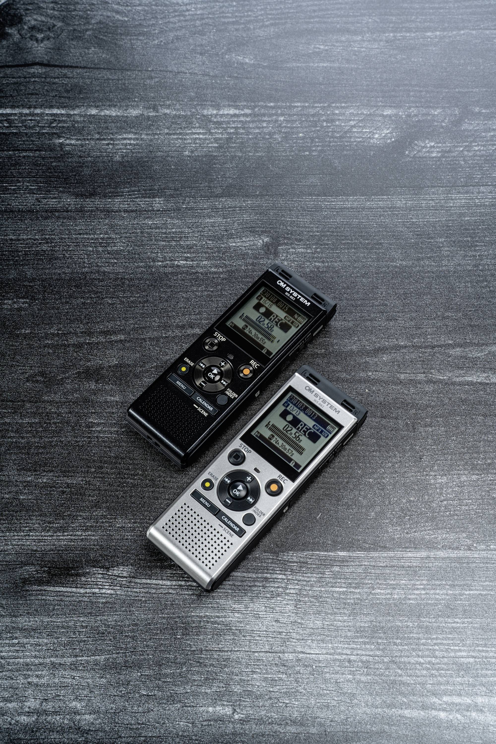 Zwei digitale Diktiergeräte liegen nebeneinander auf einem dunklen Holztisch. Eines ist schwarz, das andere silbern.