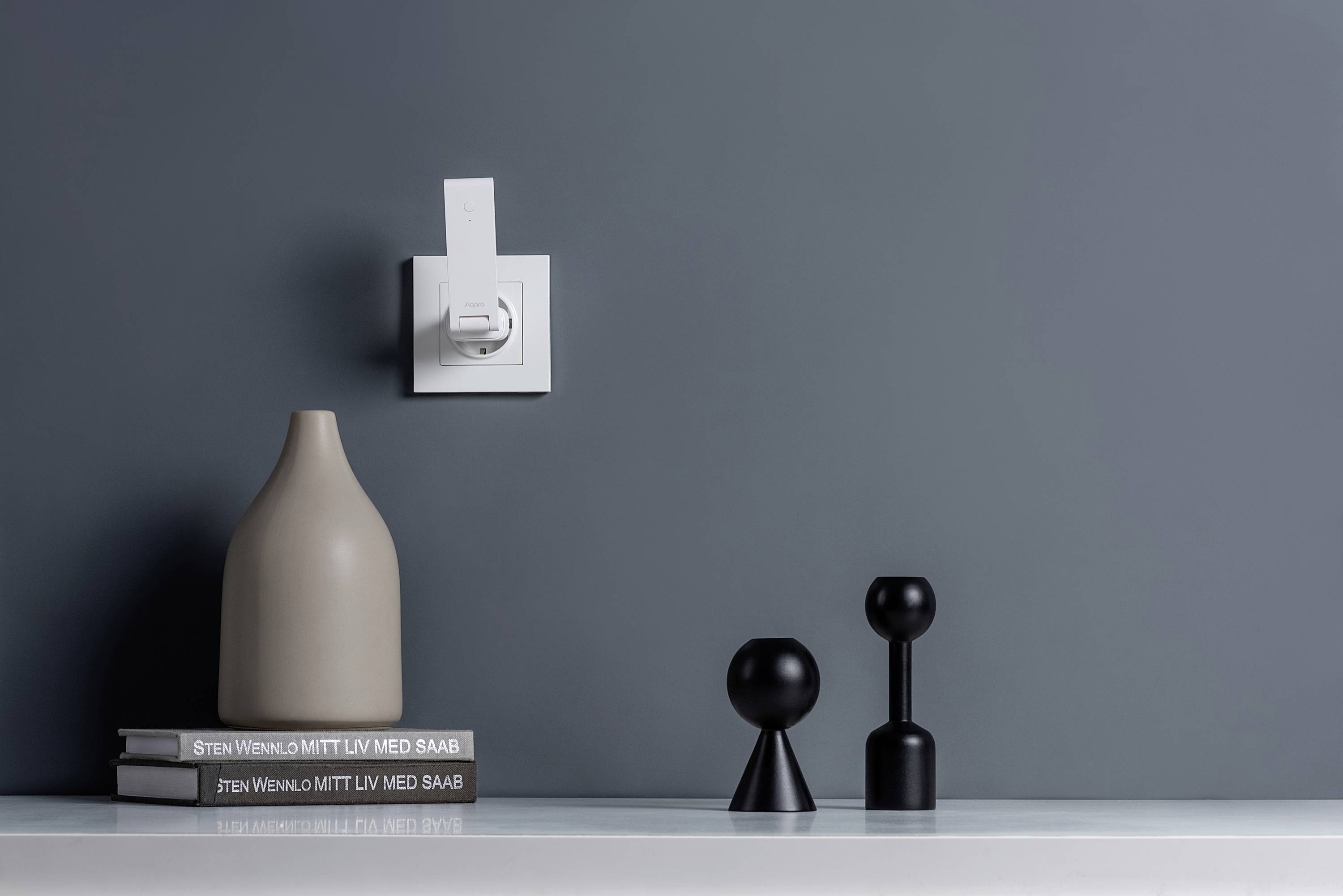 Eine moderne, minimalistische Tischdekoration mit einer grauen Vase, zwei Kerzenhaltern und drei gestapelten Büchern vor grauem Hintergrund.