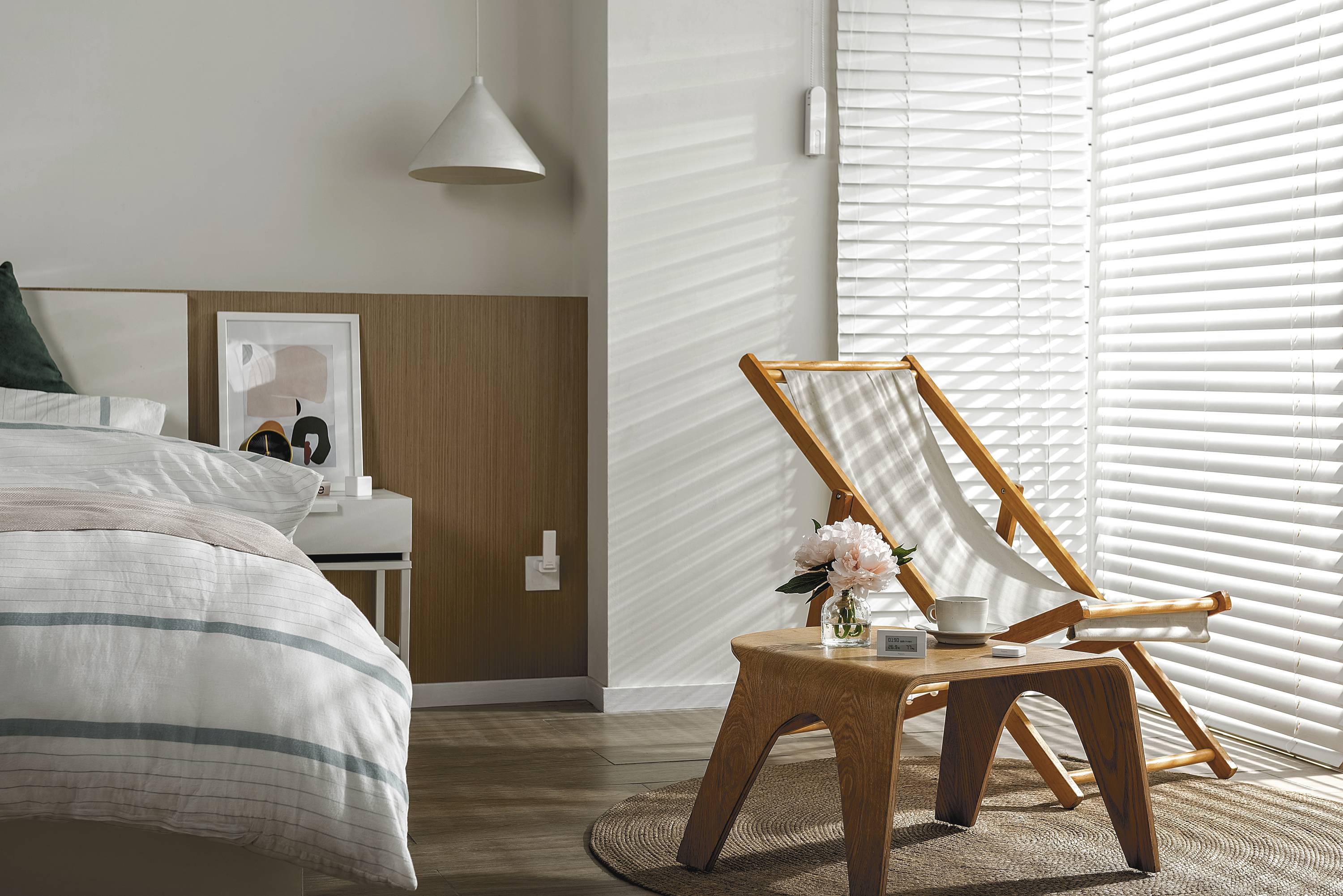 Ein helles Schlafzimmer mit einem Bett links und einem Holzstuhl neben einem kleinen Tisch auf einem runden Teppich. Sonnenlicht fällt durchs Fenster.