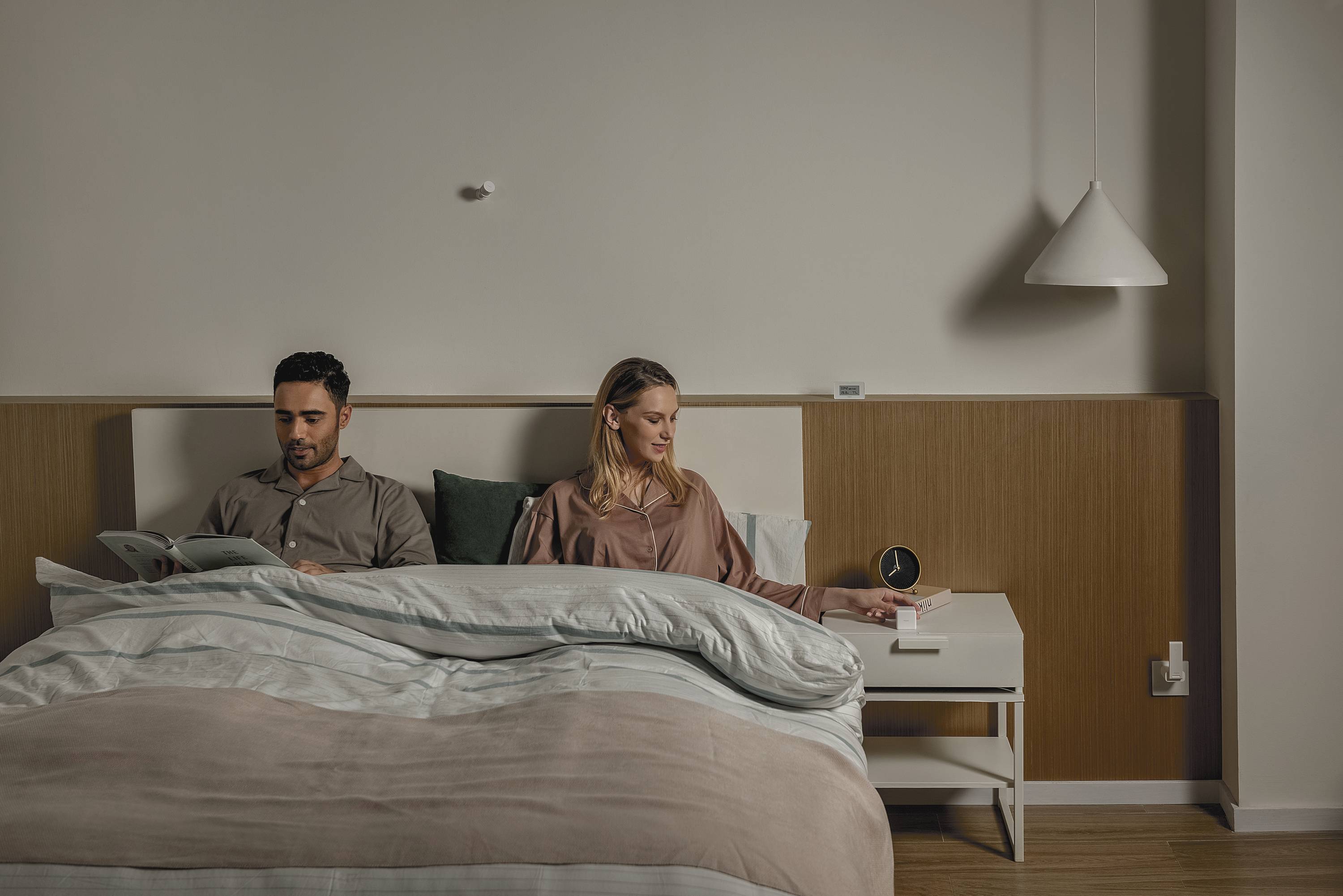 Ein Paar sitzt zusammen im Bett. Der Mann liest ein Buch, während die Frau eine Tasse greift. Sie wirken entspannt und konzentriert.