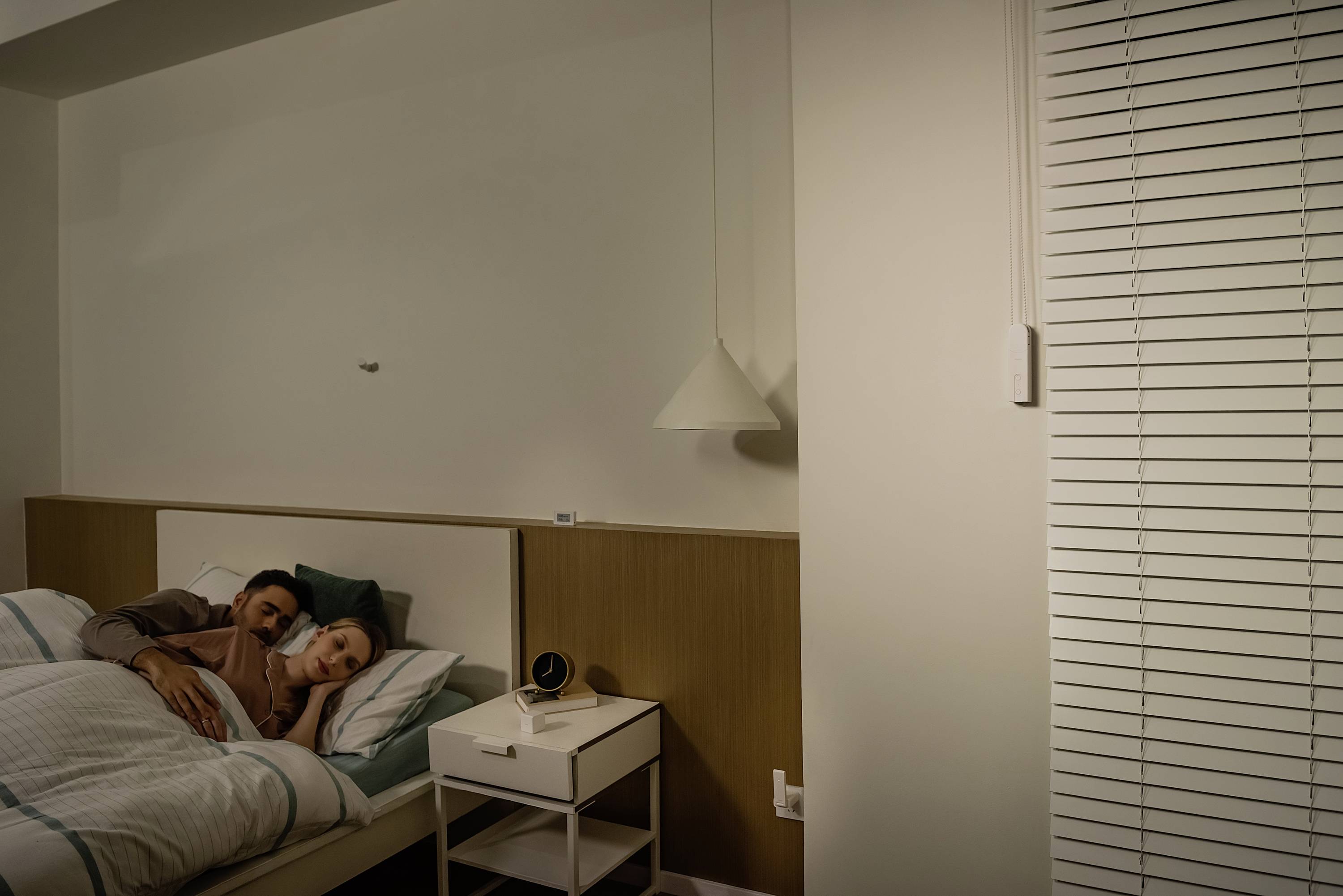 Ein Paar liegt gemütlich im Bett in einem Schlafzimmer mit gedimmter Beleuchtung. Neben dem Bett steht ein Nachttisch mit einer Lampe.