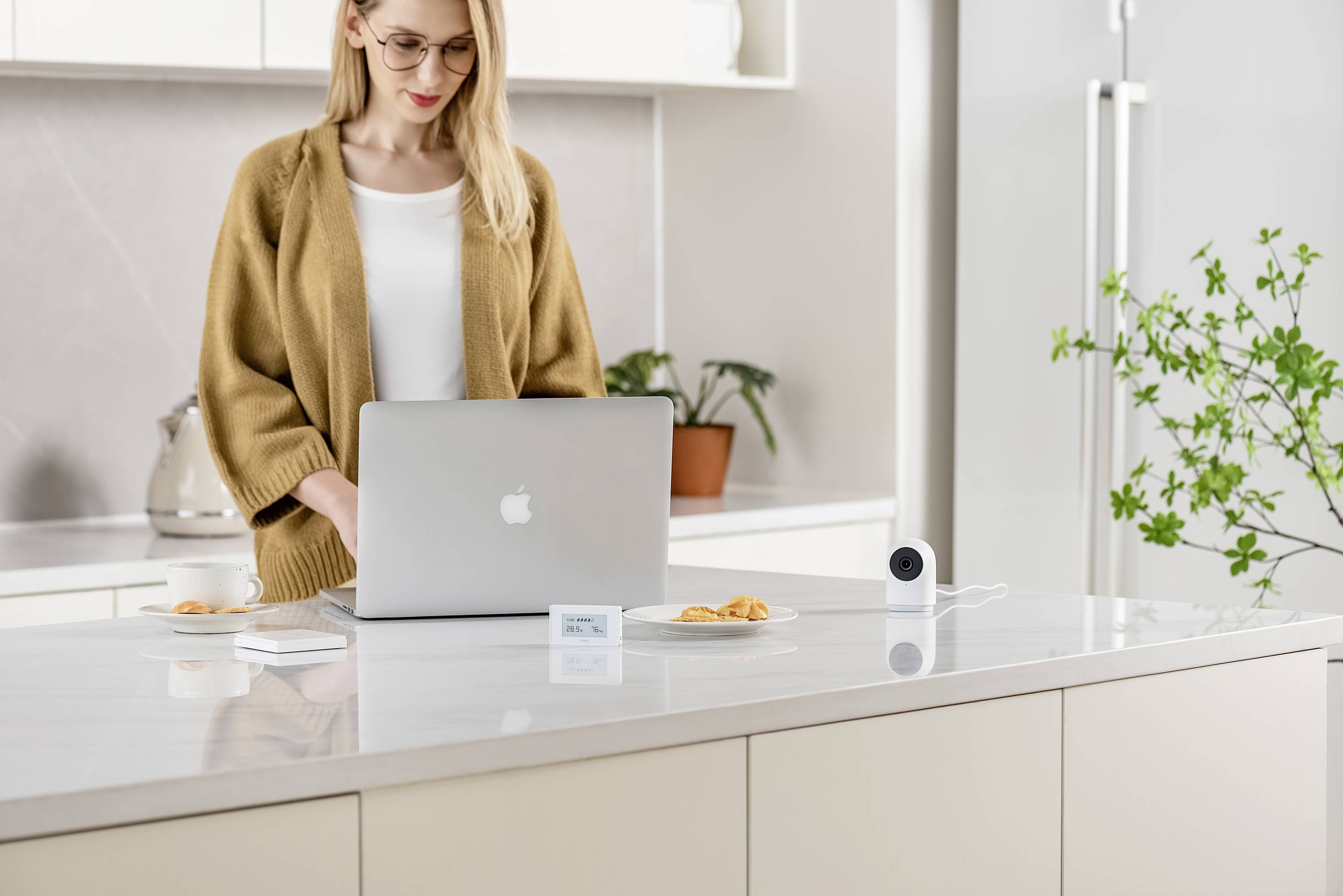 Eine Person arbeitet an einem Laptop auf einer Kücheninsel mit Croissants und einer Kamera. Im Hintergrund sind Pflanzen sichtbar.
