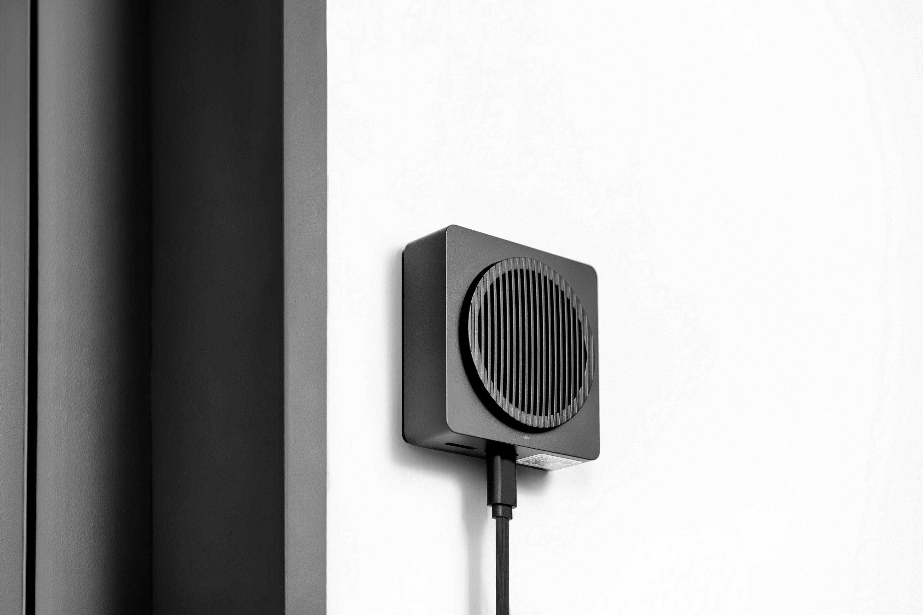 Ein kleiner, schwarzer Ventilator oder Lautsprecher ist an einer weißen Wand montiert und durch ein Kabel mit Strom versorgt.