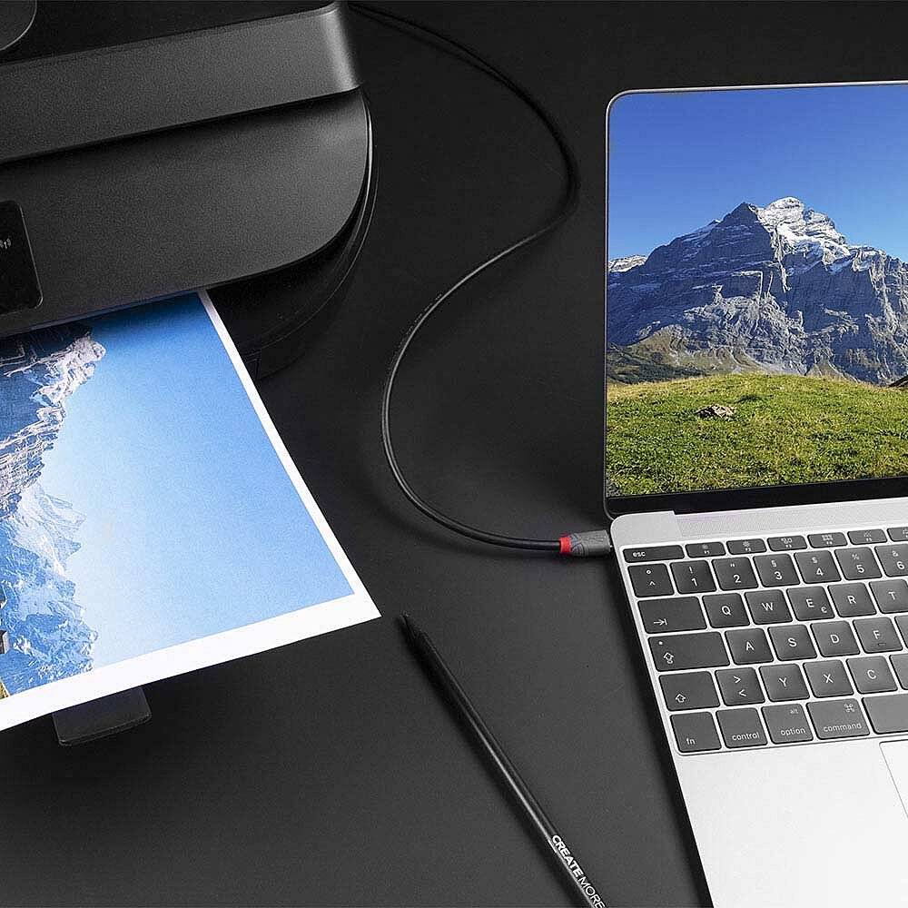 Laptop neben einem Drucker druckt ein Foto einer Berglandschaft aus. USB-Kabel verbindet Laptop und Drucker.