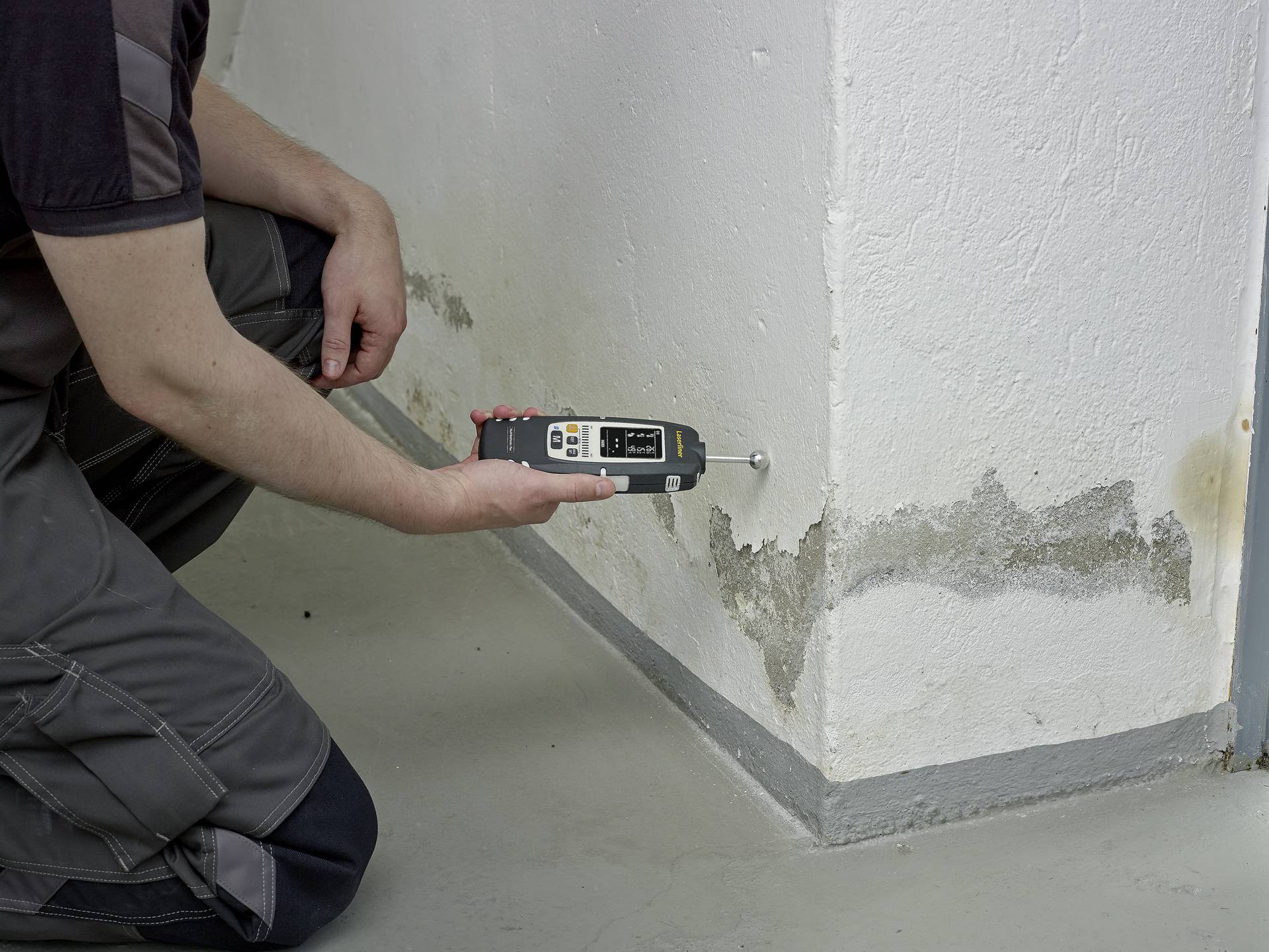 Eine Person misst mit einem Gerät die Feuchtigkeit in einer Wand, die von Wasserflecken und möglichen Schimmelschäden betroffen ist.