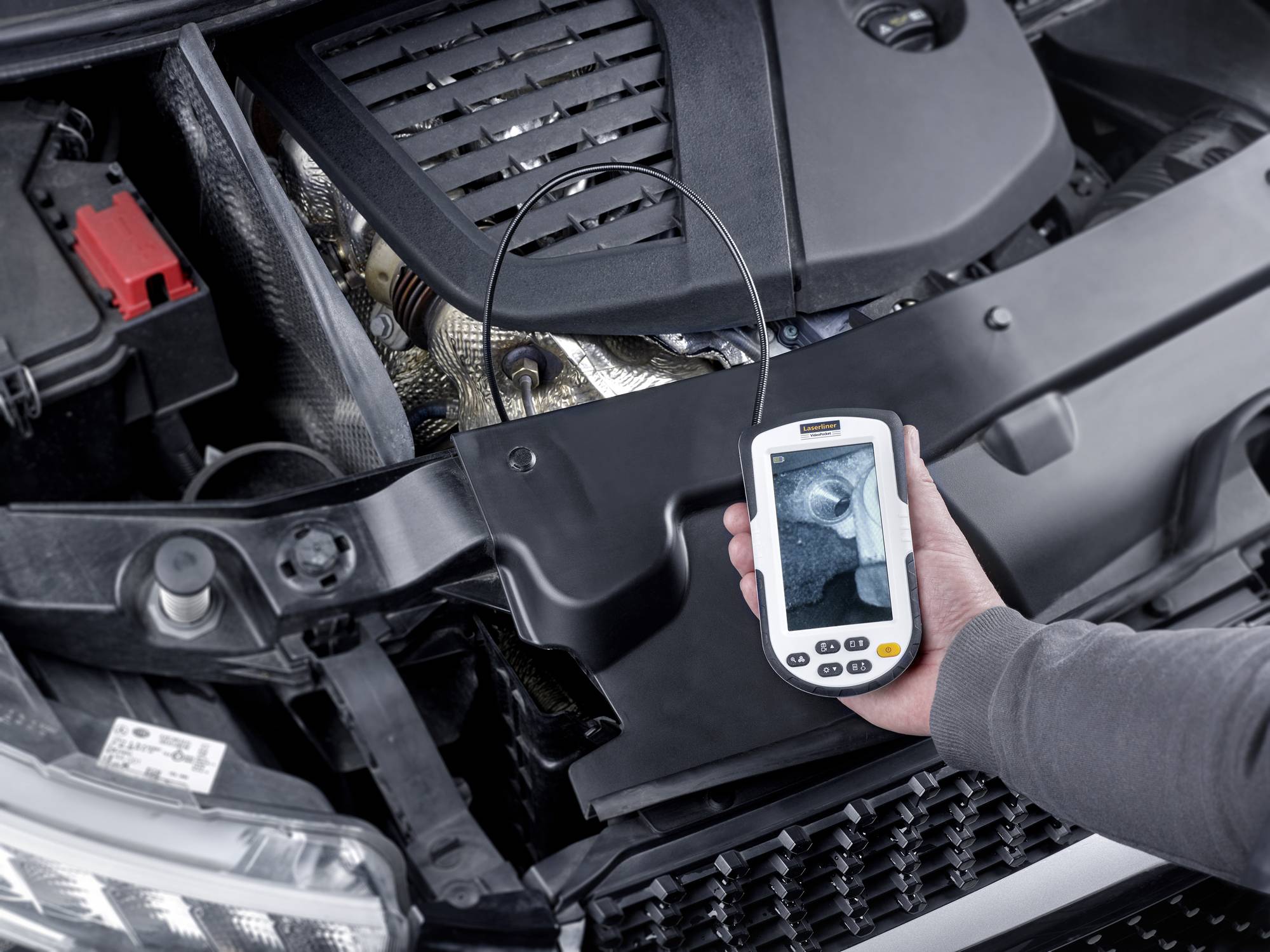 Ein Mechaniker hält ein Endoskop, das in den Motorraum eines Autos blickt. Auf dem Bildschirm ist ein vergrößertes Bild eines Motorteils zu sehen.