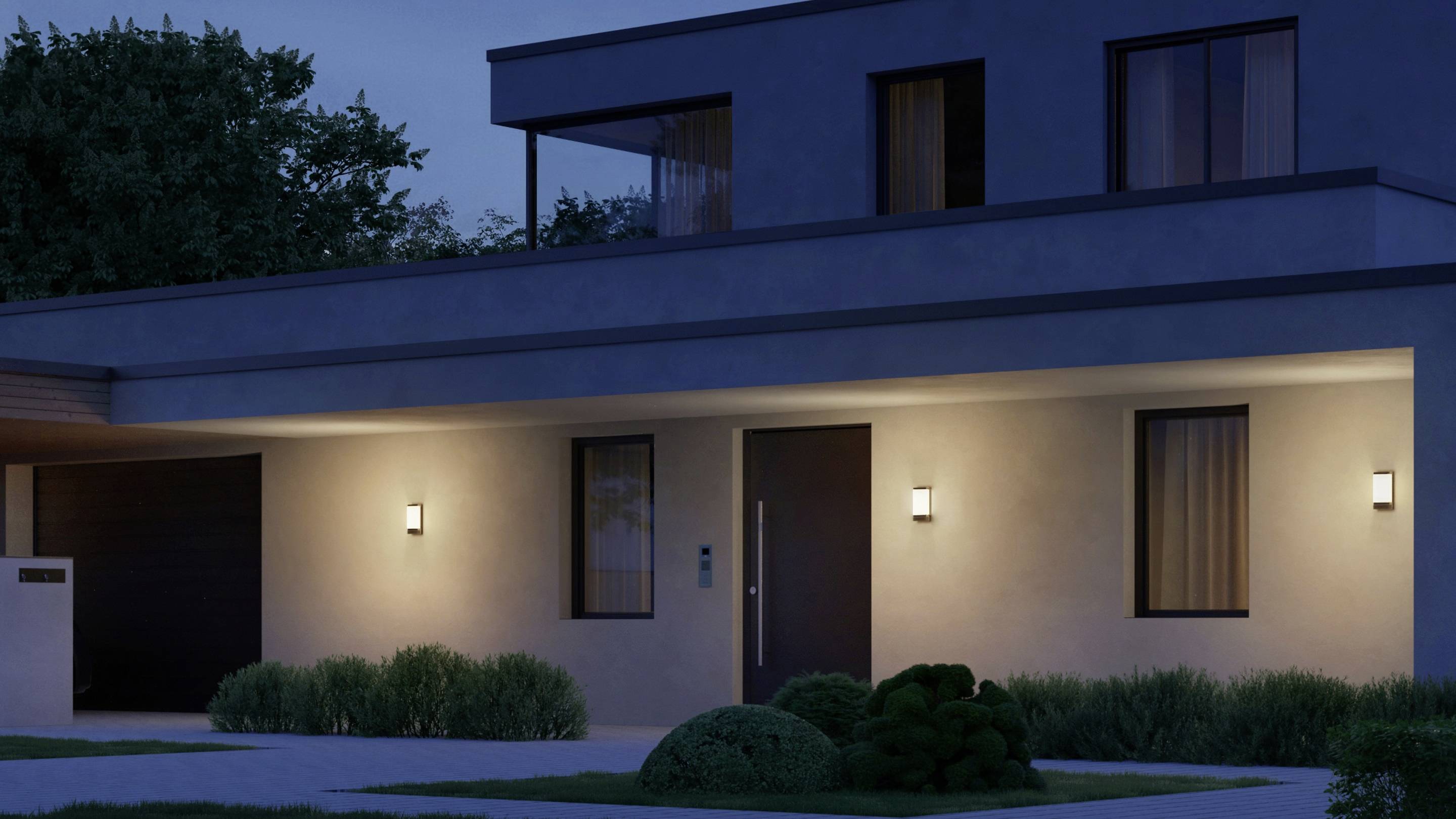 Modernes zweistöckiges Haus bei Nacht mit eingeschalteter Außenbeleuchtung. Pflanzen säumen den Gehweg vor dem Haus.