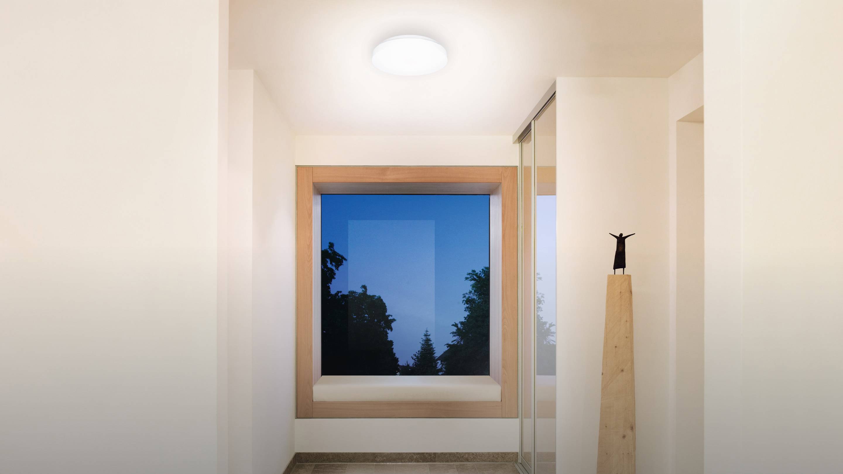 Ein schlichter, heller Flur mit großem quadratischem Fenster, das einen Ausblick auf einen blauen Himmel mit Bäumen bietet.