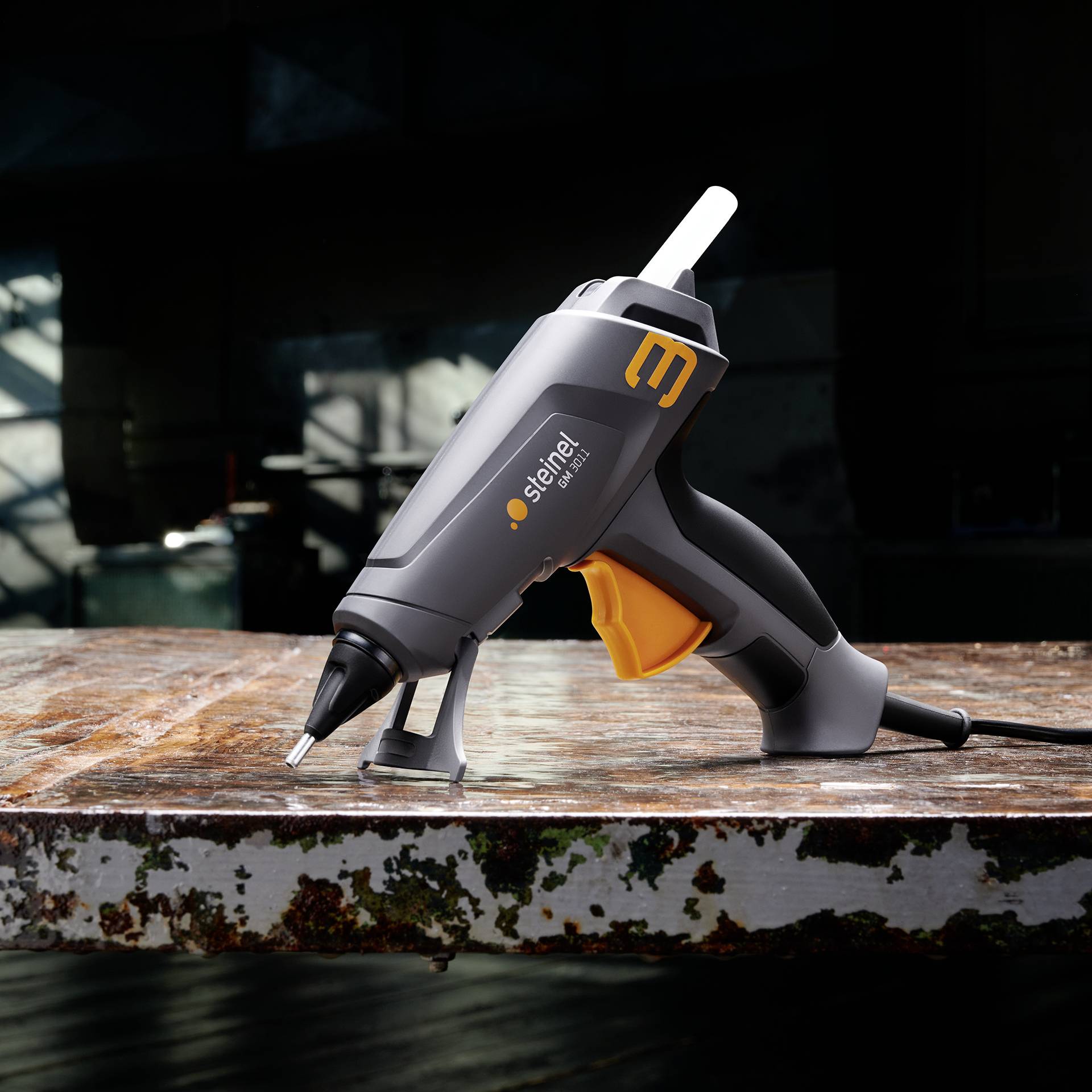 Eine schwarze Heißklebepistole liegt auf einer rostigen Metallplatte. Der Griff ist orange, und sie ist mit einem Klebestick geladen.