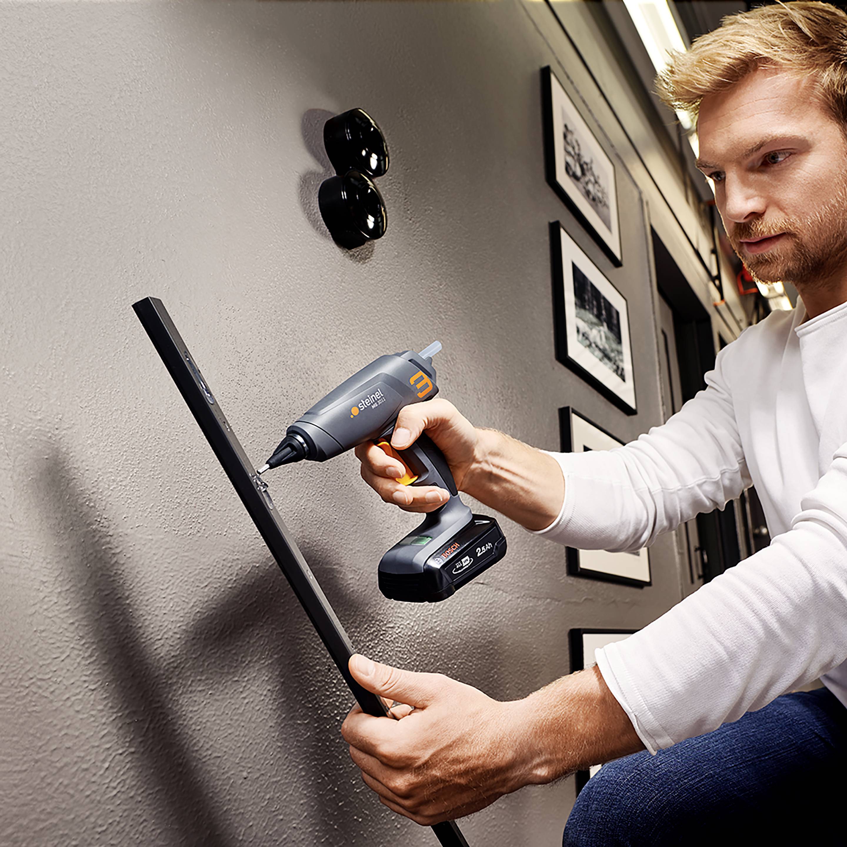 Ein Mann bohrt ein Loch in die Wand mit einem Akkuschrauber. An der Wand hängen Bilderrahmen und zwei runde Steckdosen sind sichtbar.