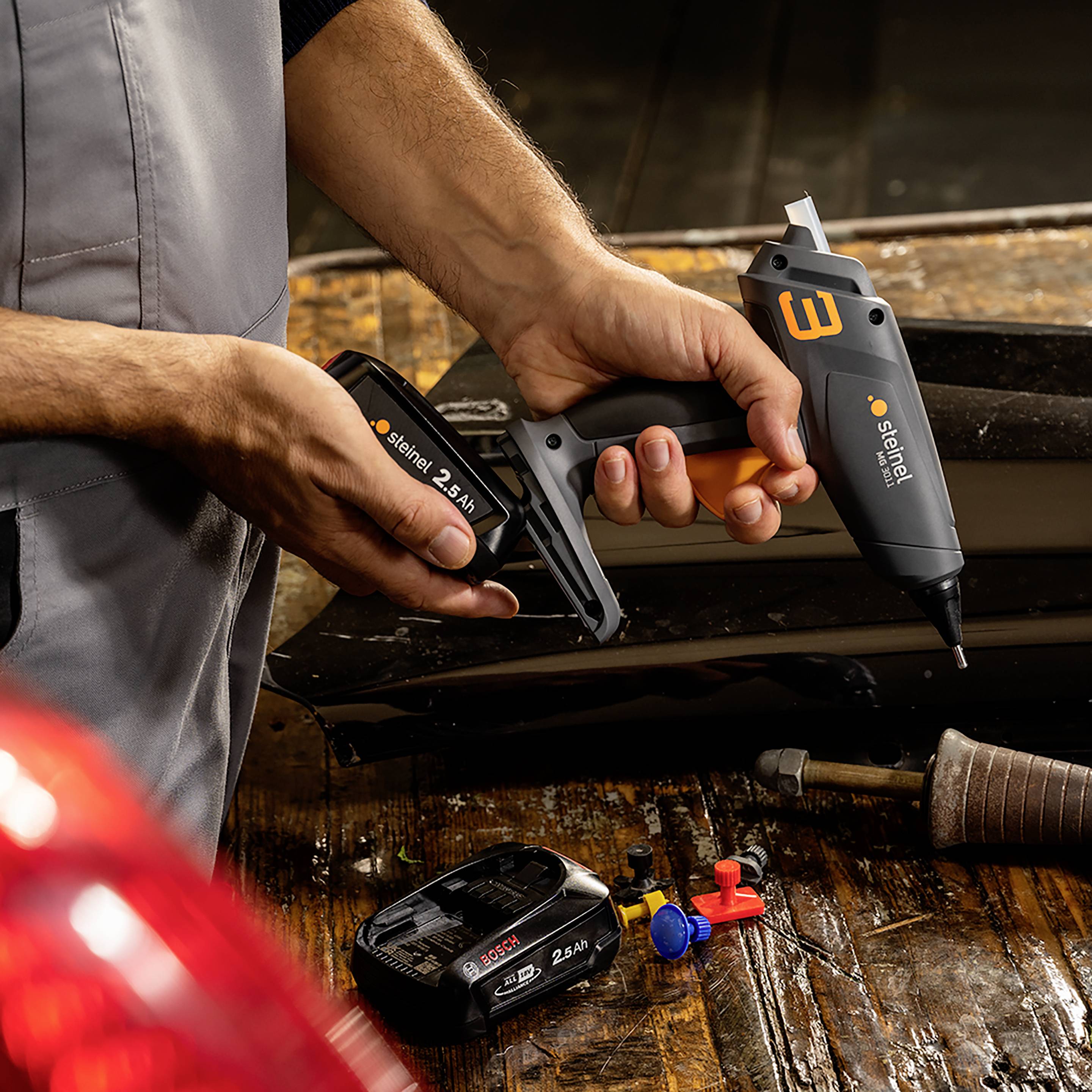 Eine Person benutzt ein elektrisches Werkzeug, um etwas auf einer Holzplatte zu befestigen. Verschiedene Dübel und eine Batterie liegen daneben.