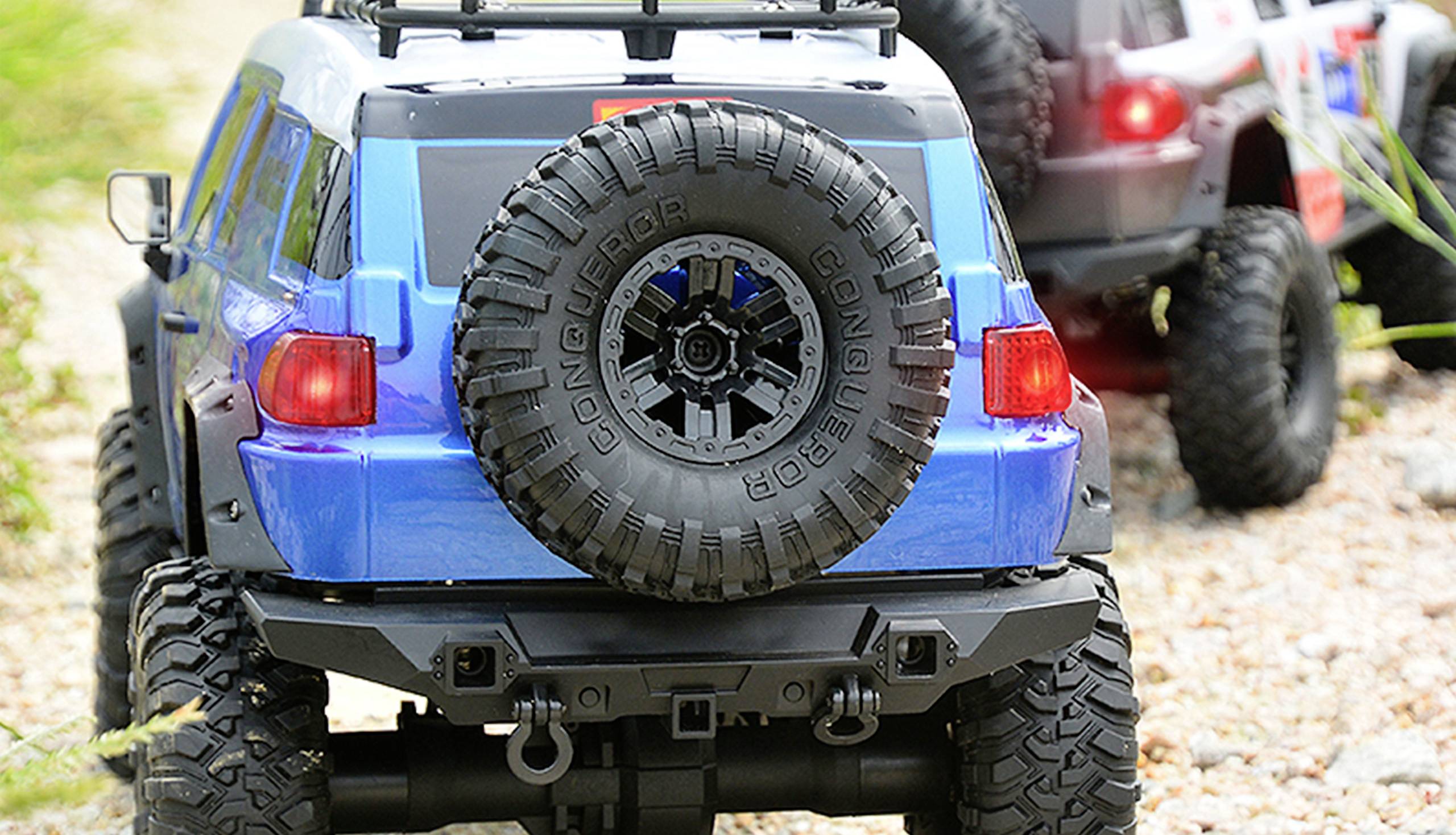 Zwei ferngesteuerte Geländewagen auf einem steinigen Pfad. Der vordere ist blau und trägt ein Reserverad mit der Aufschrift 'Conqueror'.