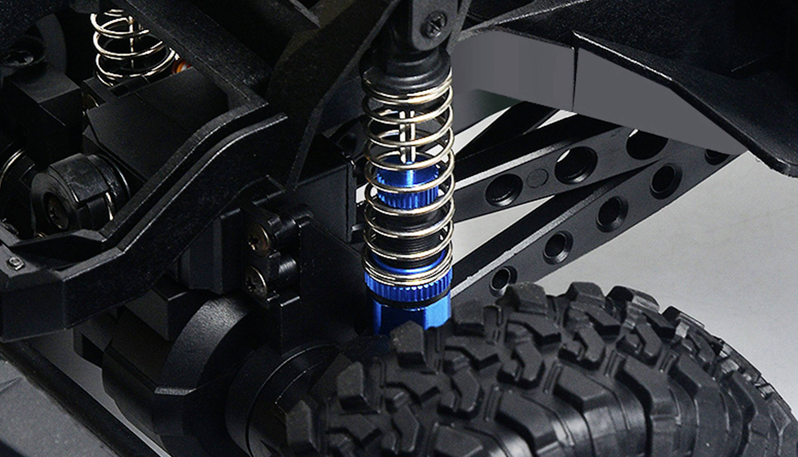 Nahaufnahme einer Fahrzeugfederung mit blauer Stoßdämpferfeder über einem grobstolligen Reifen. Mechanische Details im Hintergrund.