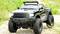 Ferngesteuertes schwarzes Offroad-Modellauto mit groben Reifen fährt auf einem steinigen Weg, umgeben von Gras.