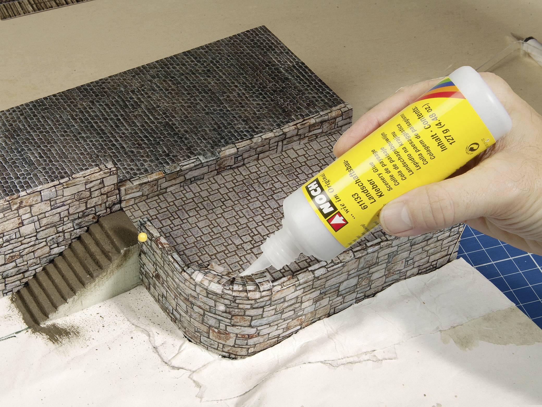 Eine Person klebt detailreiche Ziegel auf ein Miniaturmodell einer Gebäudewand mit einer Klebeflasche.