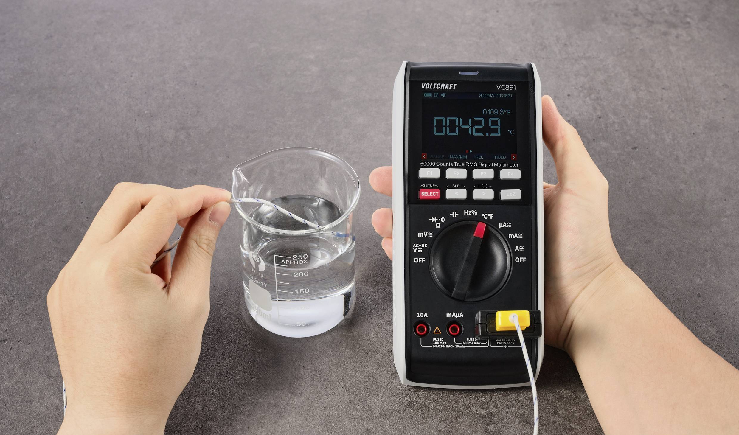Eine Hand hält ein Multimeter, das mit einem Thermoelement verbunden ist. Eine andere Hand taucht das Thermoelement in ein Glas Wasser. Das Display zeigt '00.42.9°C'.