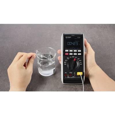 Eine Hand hält ein Multimeter, das mit einem Thermoelement verbunden ist. Eine andere Hand taucht das Thermoelement in ein Glas Wasser. Das Display zeigt '00.42.9°C'.