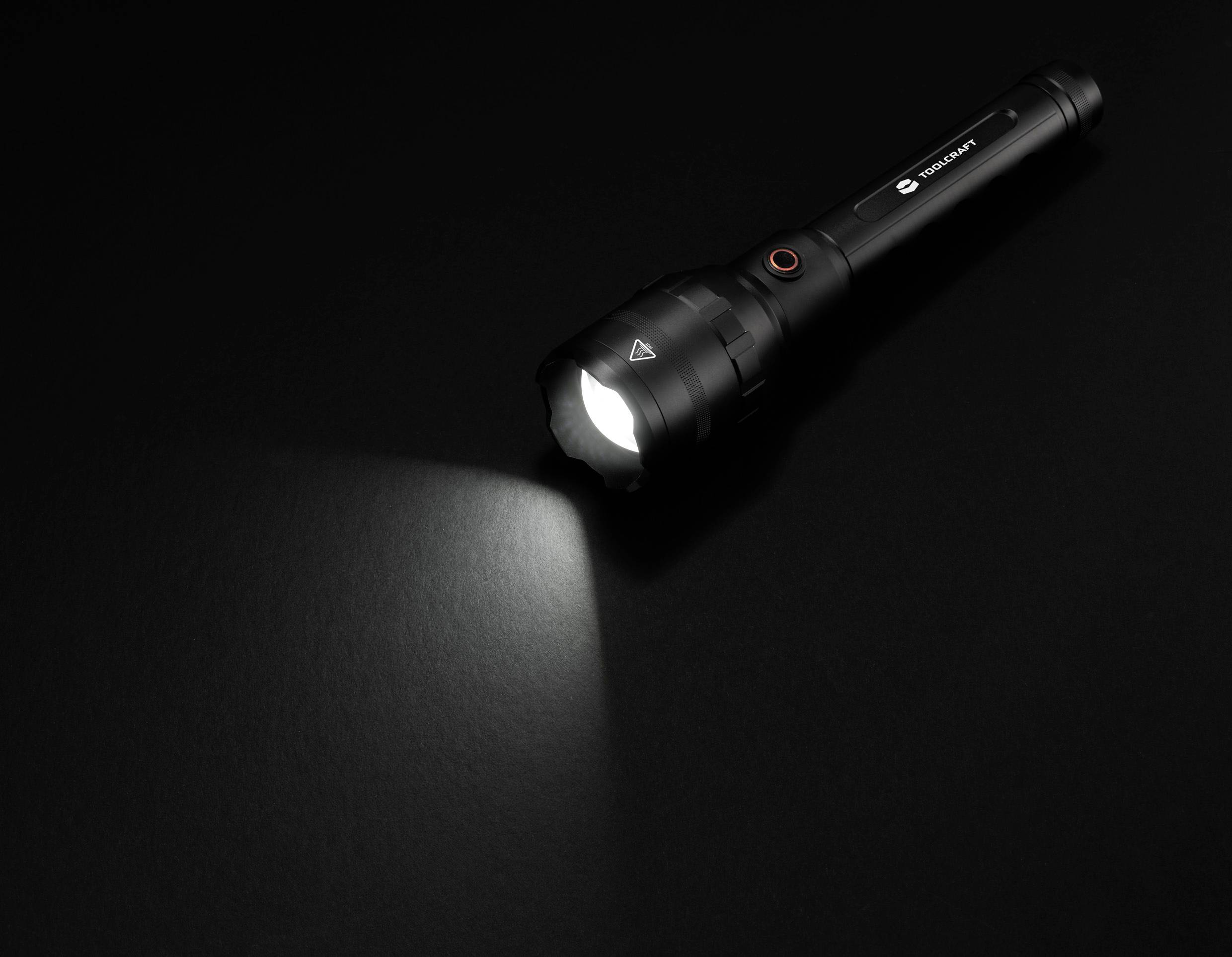 Eine schwarze Taschenlampe liegt auf einem dunklen Hintergrund. Sie ist eingeschaltet und wirft einen Lichtstrahl nach links.