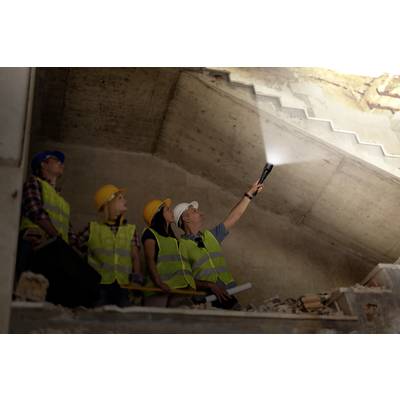 'Arbeiter inspizieren eine Baustelle in einem Rohbau. Eine Person leuchtet mit einer Taschenlampe an die Decke. Alle tragen Schutzhelme und Warnwesten.'