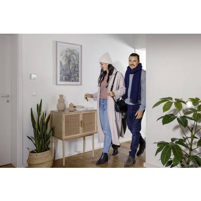 Ein Mann und eine Frau betreten lächelnd einen Raum, beide in Winterkleidung. Im Hintergrund sind Pflanzen und ein Schrank zu sehen.