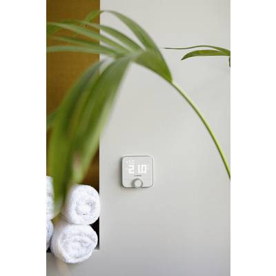 Ein digitaler Thermostat zeigt Temperatur an. Im Vordergrund ein Blatt einer Zimmerpflanze, daneben aufgerollte Handtücher.