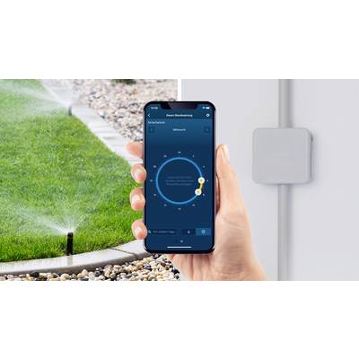 Eine Hand hält ein Smartphone mit geöffneter Bewässerungs-App. Im Hintergrund bewässert ein Sprinkler den Rasen.
