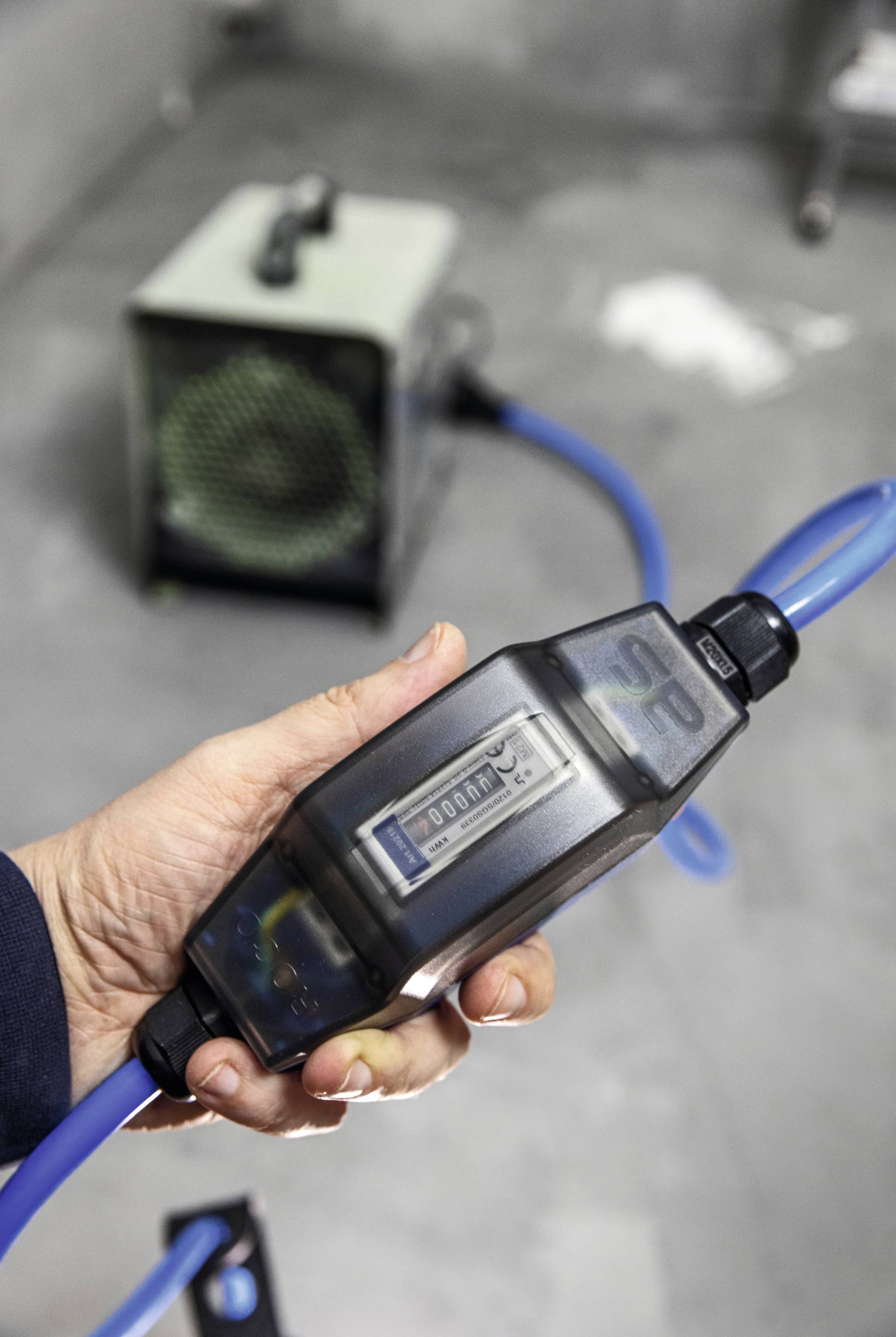 Eine Hand hält ein digitales Messgerät, das an einen blauen Schlauch angeschlossen ist. Im Hintergrund ist ein weiteres Gerät zu sehen.