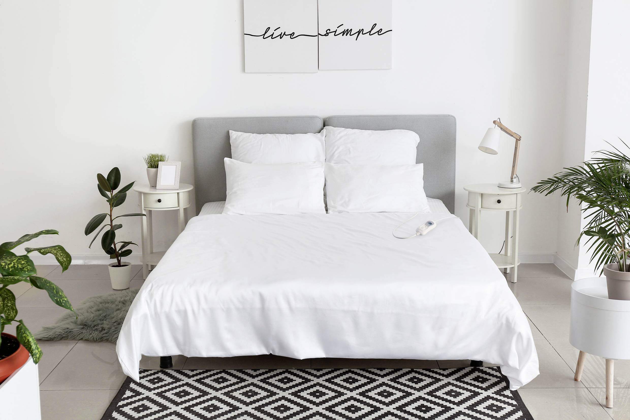 Ein gemütliches Schlafzimmer mit einem grauen Bett, umgeben von Pflanzen. An der Wand hängt ein Bild mit dem Text 'live simple'.