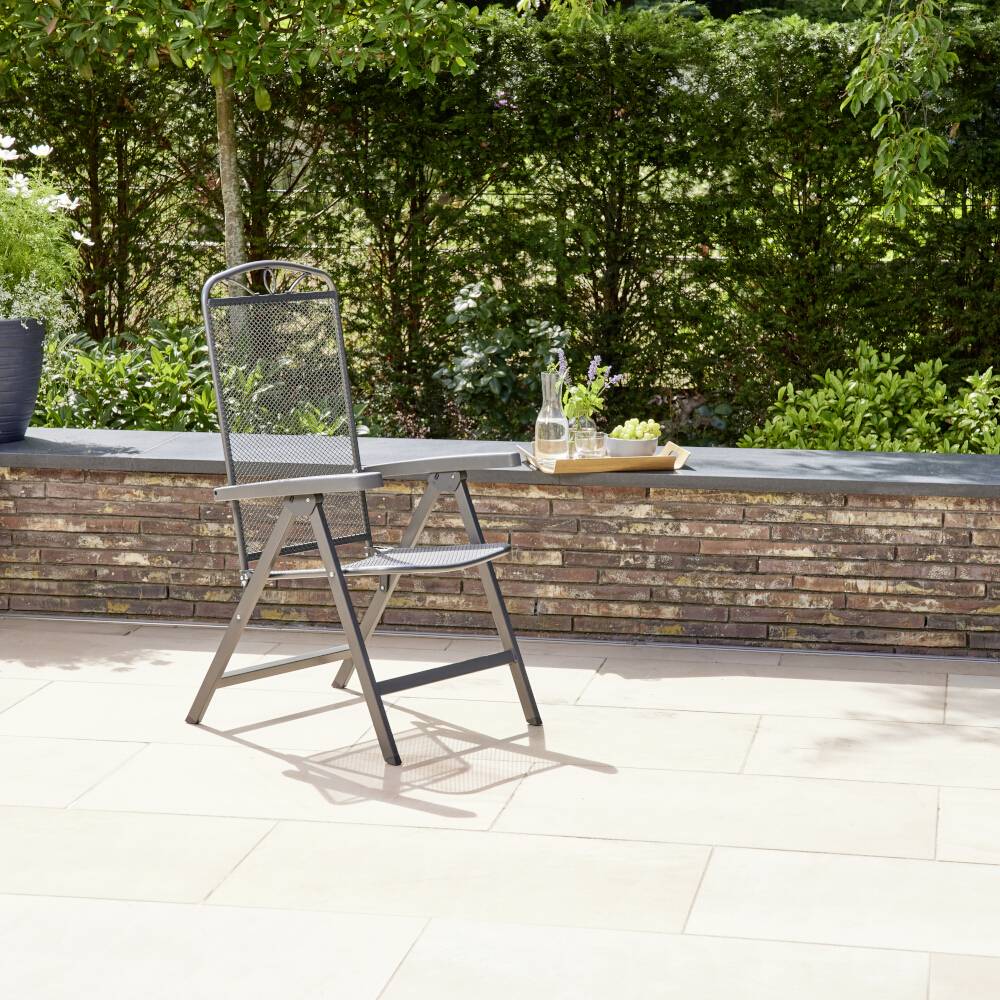 Ein metallener Gartenstuhl steht auf einer hellen Terrasse vor einer niedrigen Backsteinmauer, flankiert von Pflanzen und Büschen.