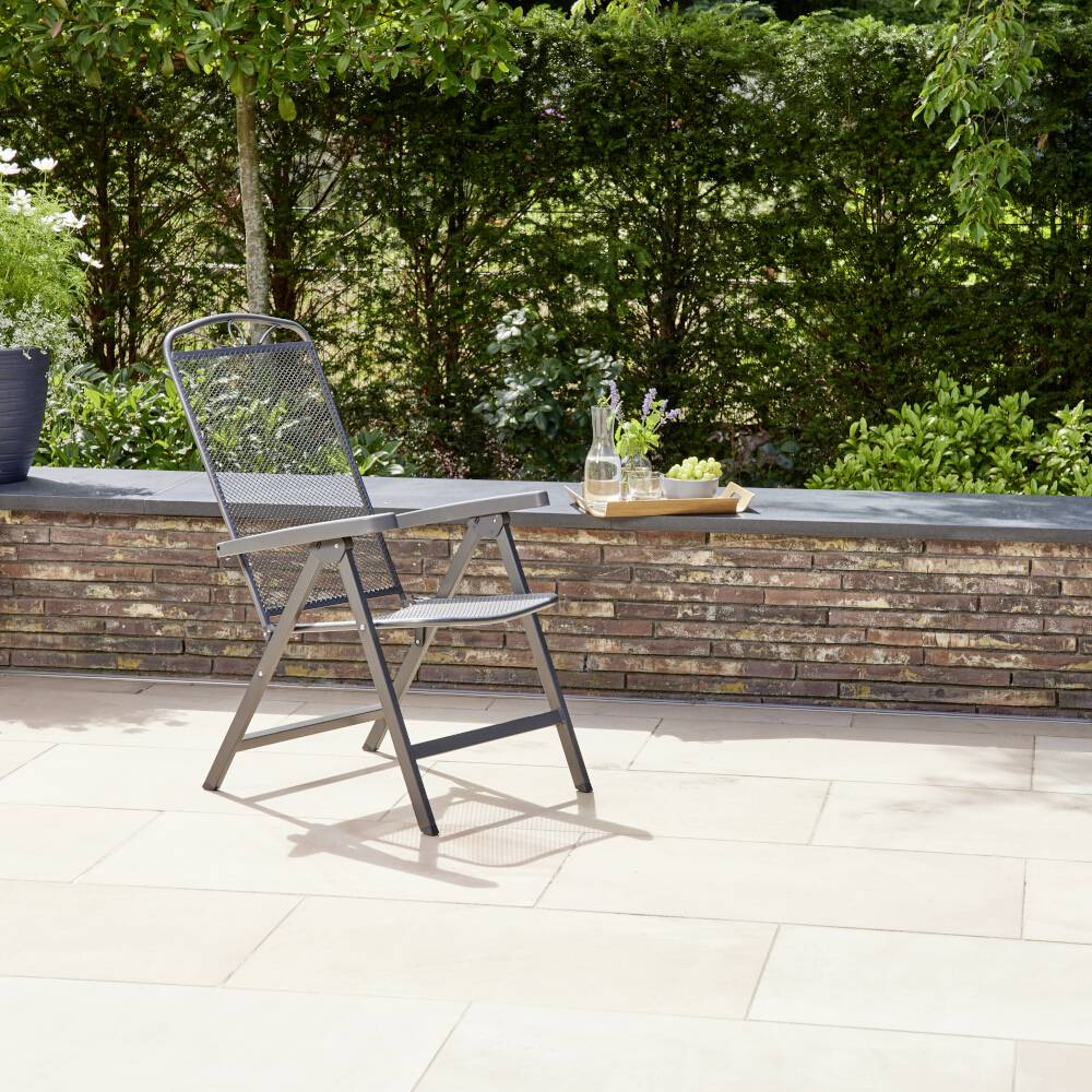 Ein einzelner Metallstuhl steht auf einer Terrasse mit hellem Steinboden. Im Hintergrund sind ein niedriger Ziegelsteinzaun und grüne Büsche zu sehen.