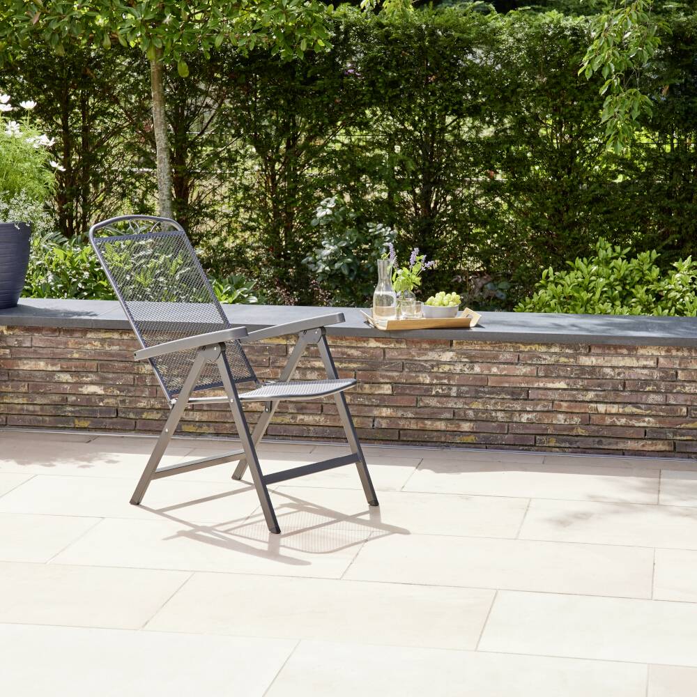Ein einzelner Metallstuhl steht auf einer Terrasse aus hellen Fliesen, umgeben von grüner Vegetation und einer gemauerten Mauer.