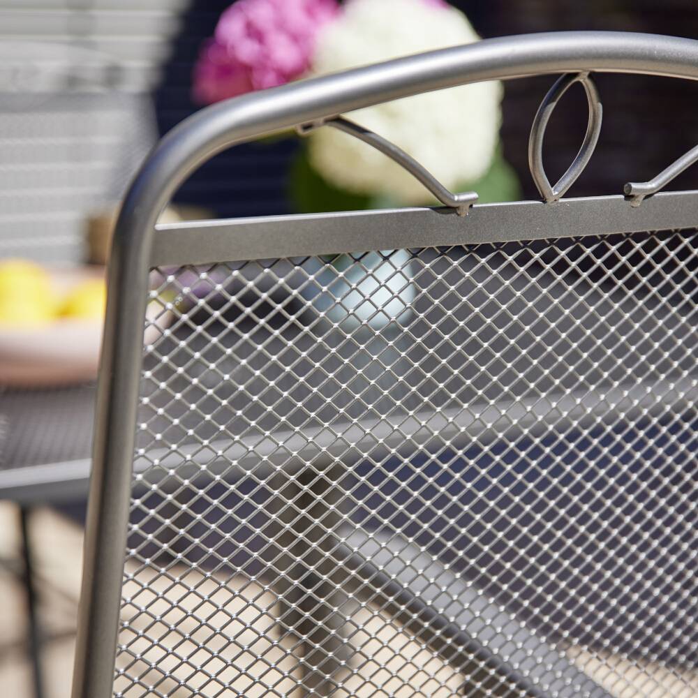 Ein Metallstuhl im Vordergrund mit unscharfen, rosa und weißen Blumen im Hintergrund auf einer Terrasse.