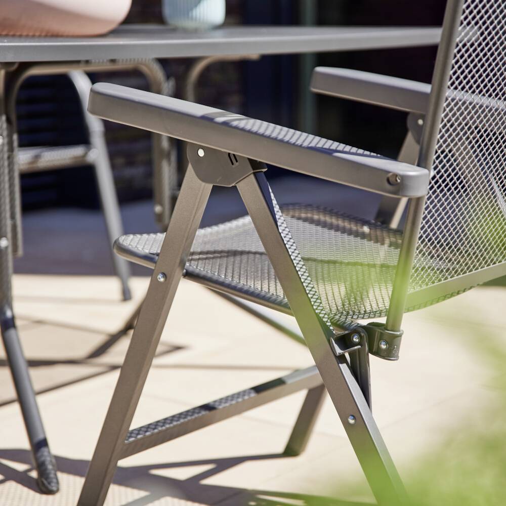 Ein Metallstuhl steht auf einer Terrasse, teilweise von grünen Blättern verdeckt. Im Hintergrund ist ein Tisch erkennbar.