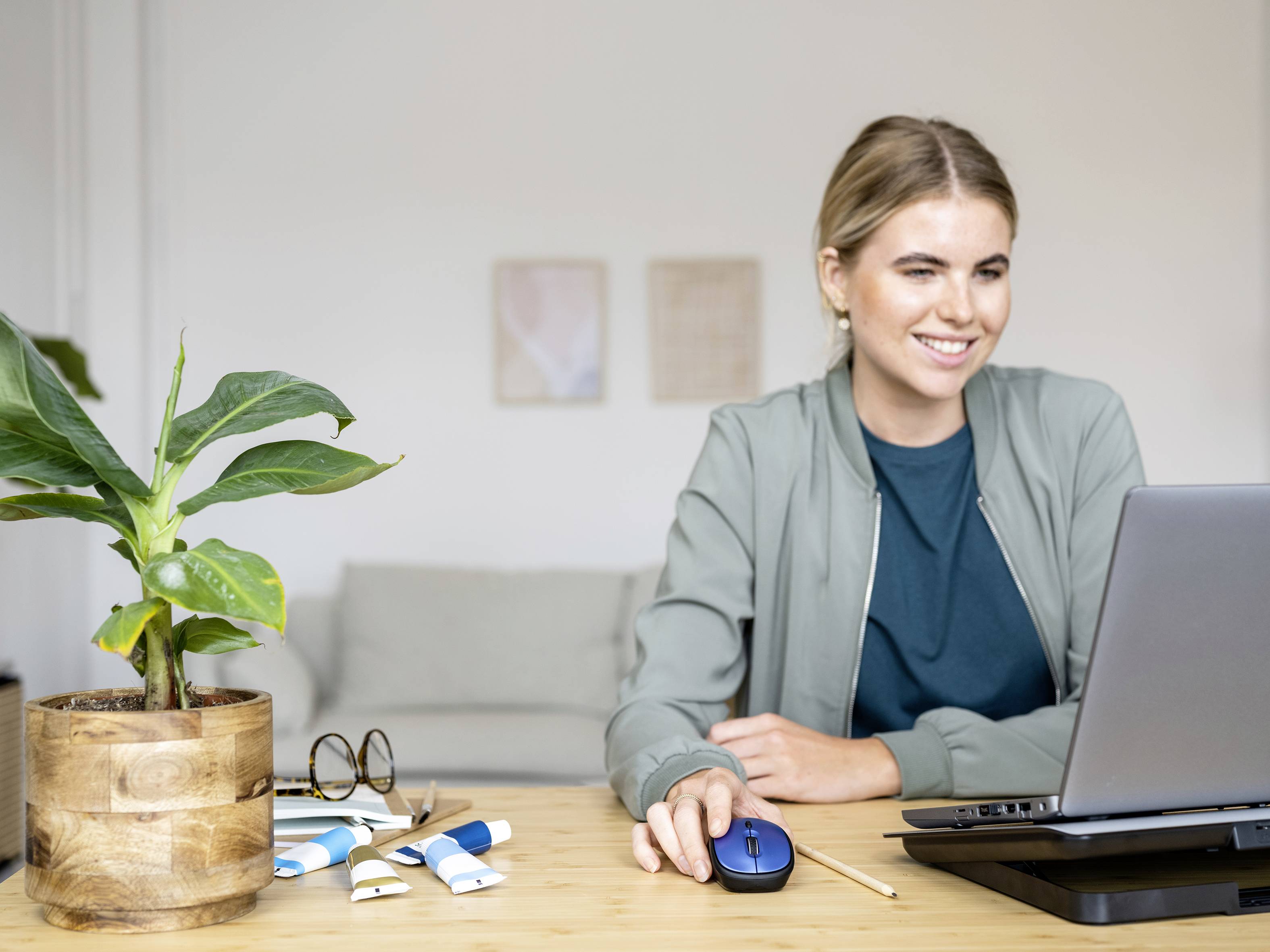 Frau sitzt lächelnd vor einem Laptop an einem Tisch mit Pflanze und Büroartikeln.