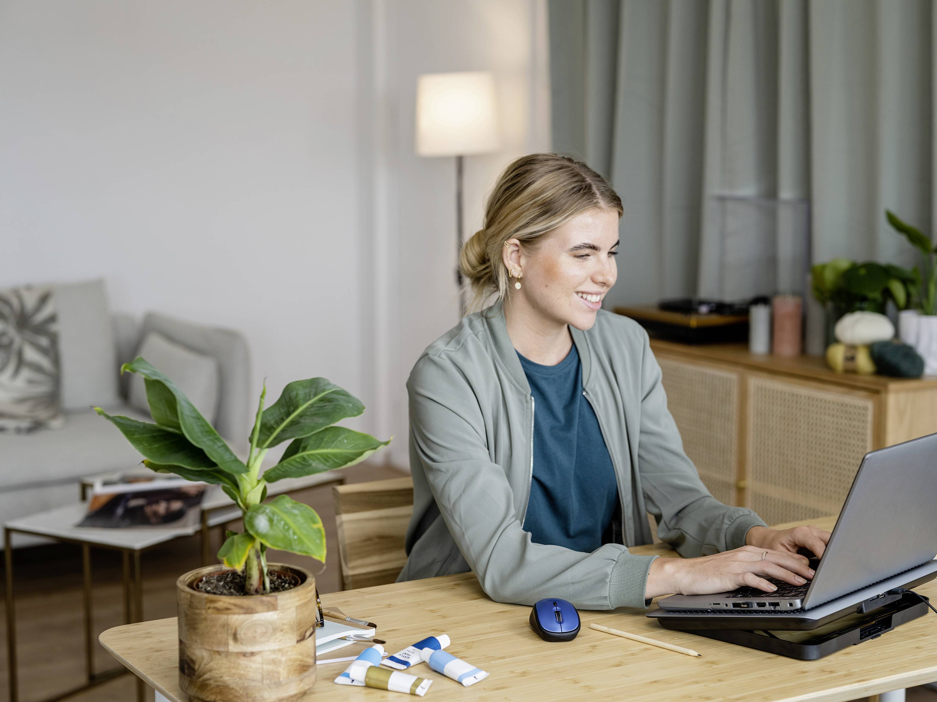 Frau mit blonden Haaren sitzt am Schreibtisch und arbeitet lächelnd an einem Laptop; Topfpflanze und Farbtuben im Vordergrund.